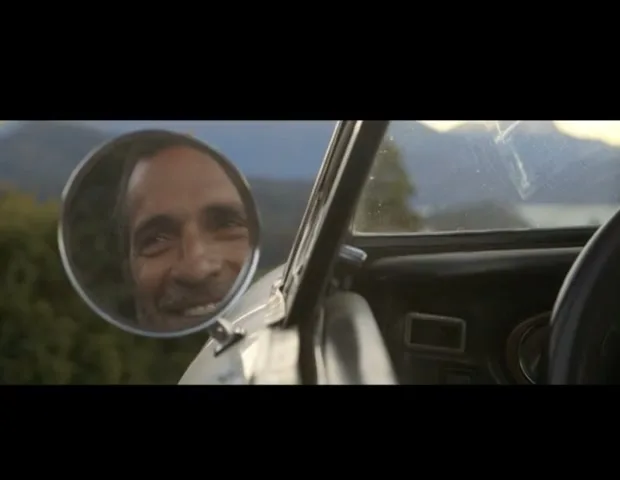 A man with a beard is seen smiling in a car's side mirror, with a green landscape and mountains in the background.