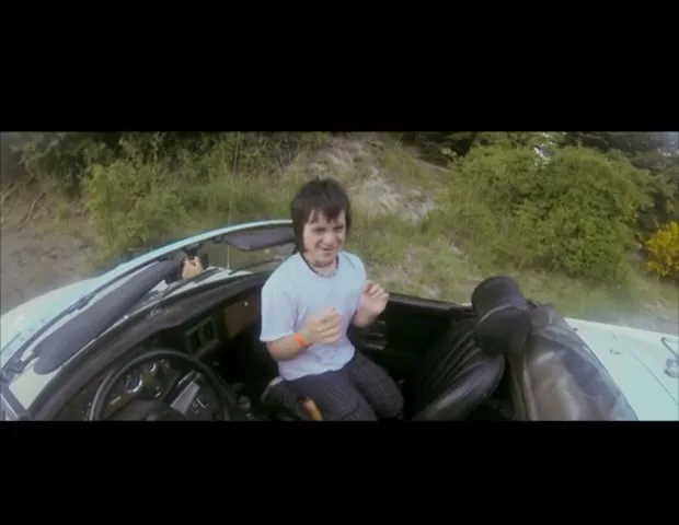 A young man with dark hair and a white t-shirt sits in the passenger seat of a white convertible, looking scared and gesturing with his hands, on a dirt road surrounded by green foliage.