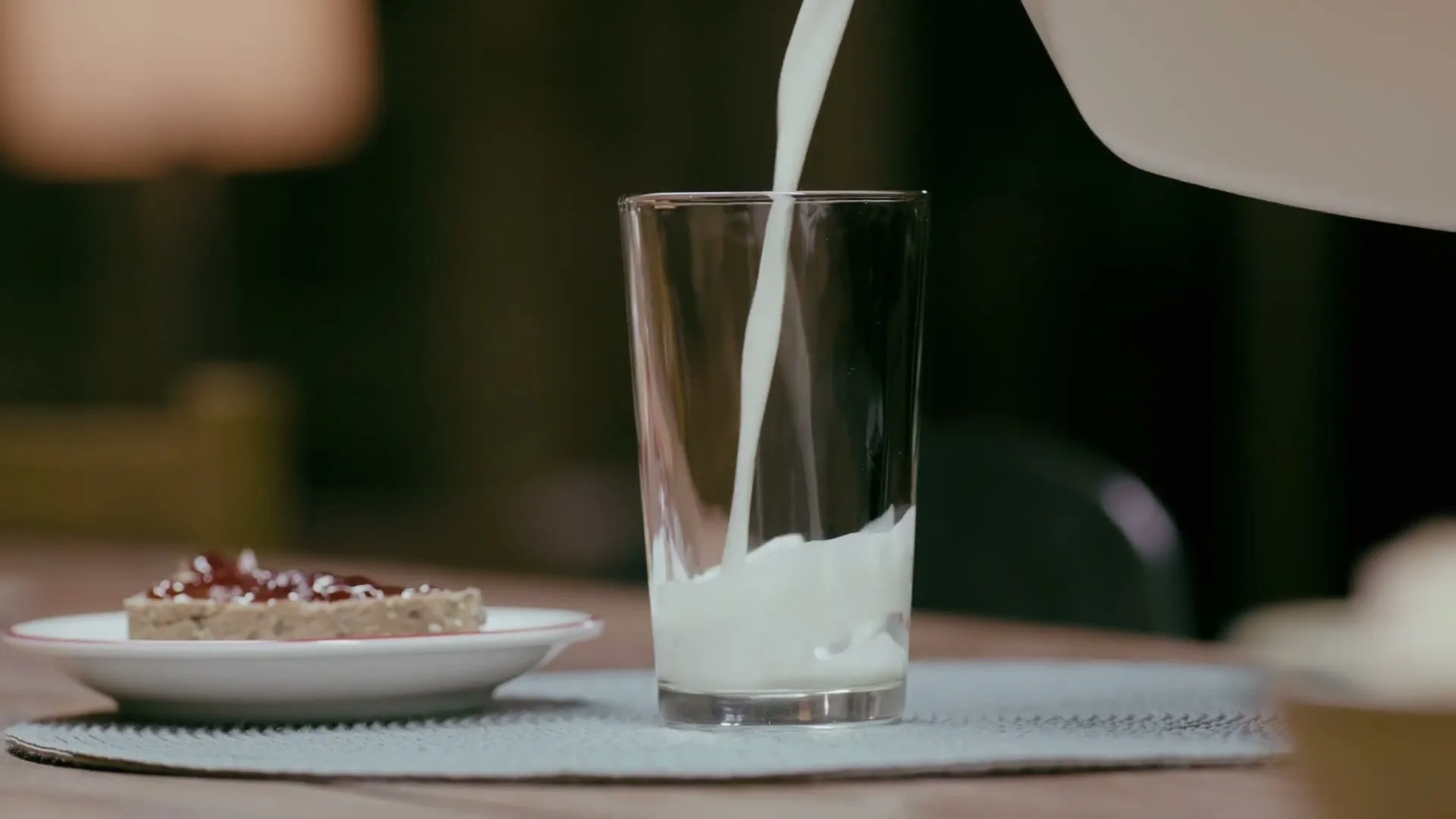 Milk is poured from a white pitcher into a clear glass on a table next to a piece of toast with jam.