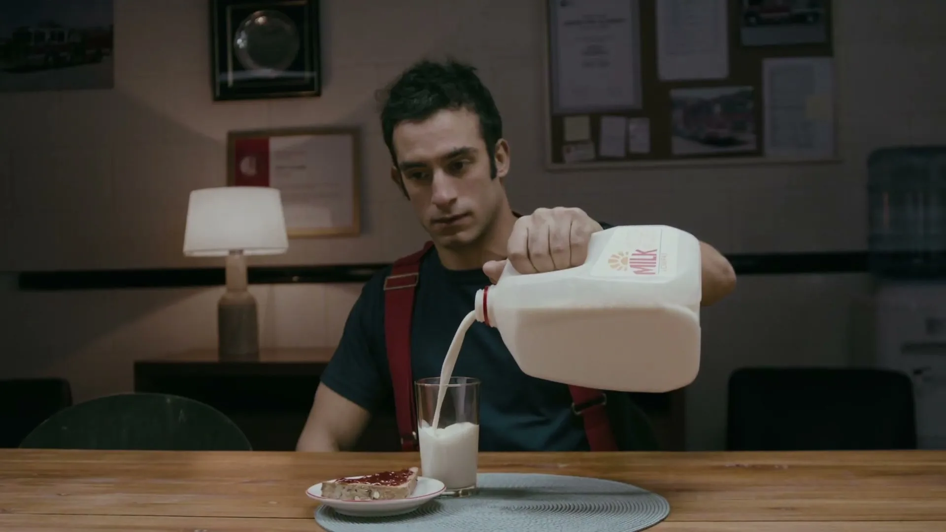 A young male firefighter in a dark t-shirt and red suspenders pours milk from a gallon jug into a glass at a wooden table in a fire station.
