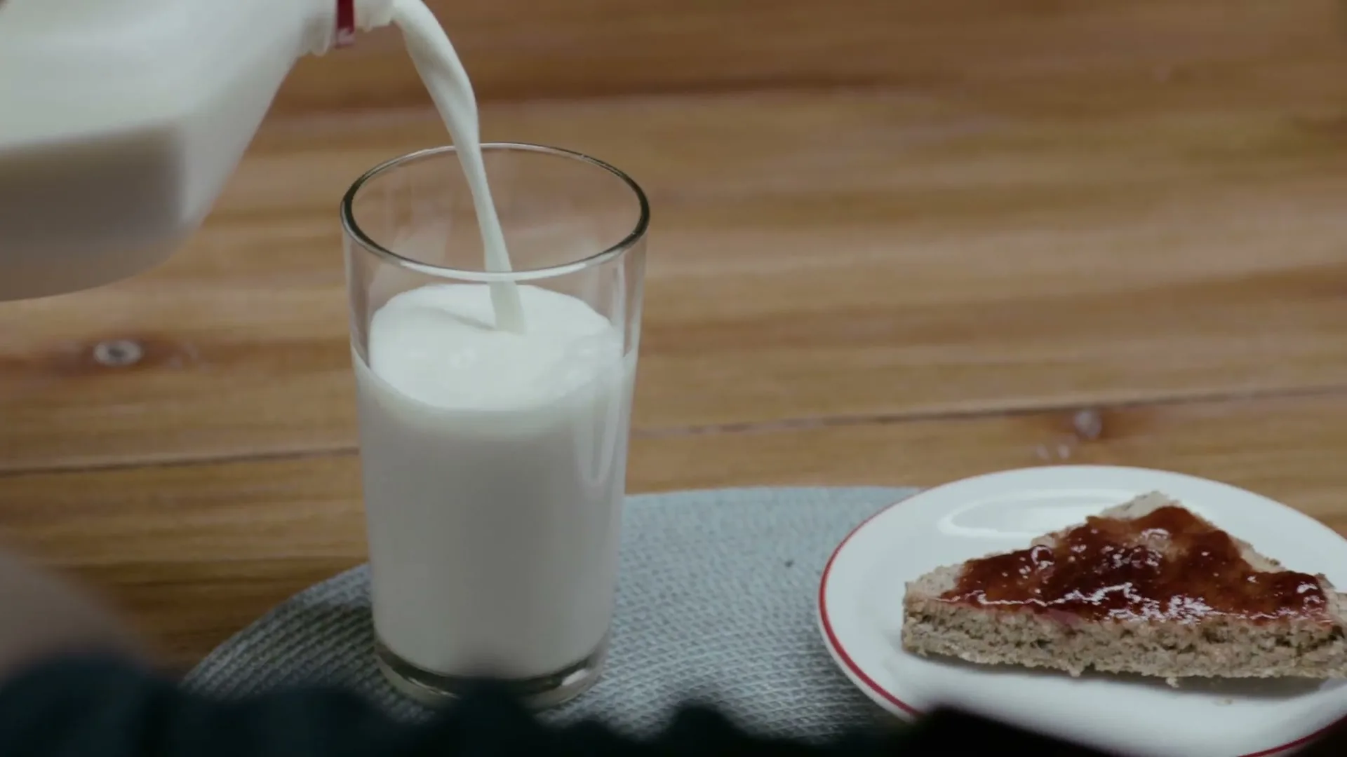 Milk is poured from a white carton into a glass next to a piece of toast with jam on a wooden table.