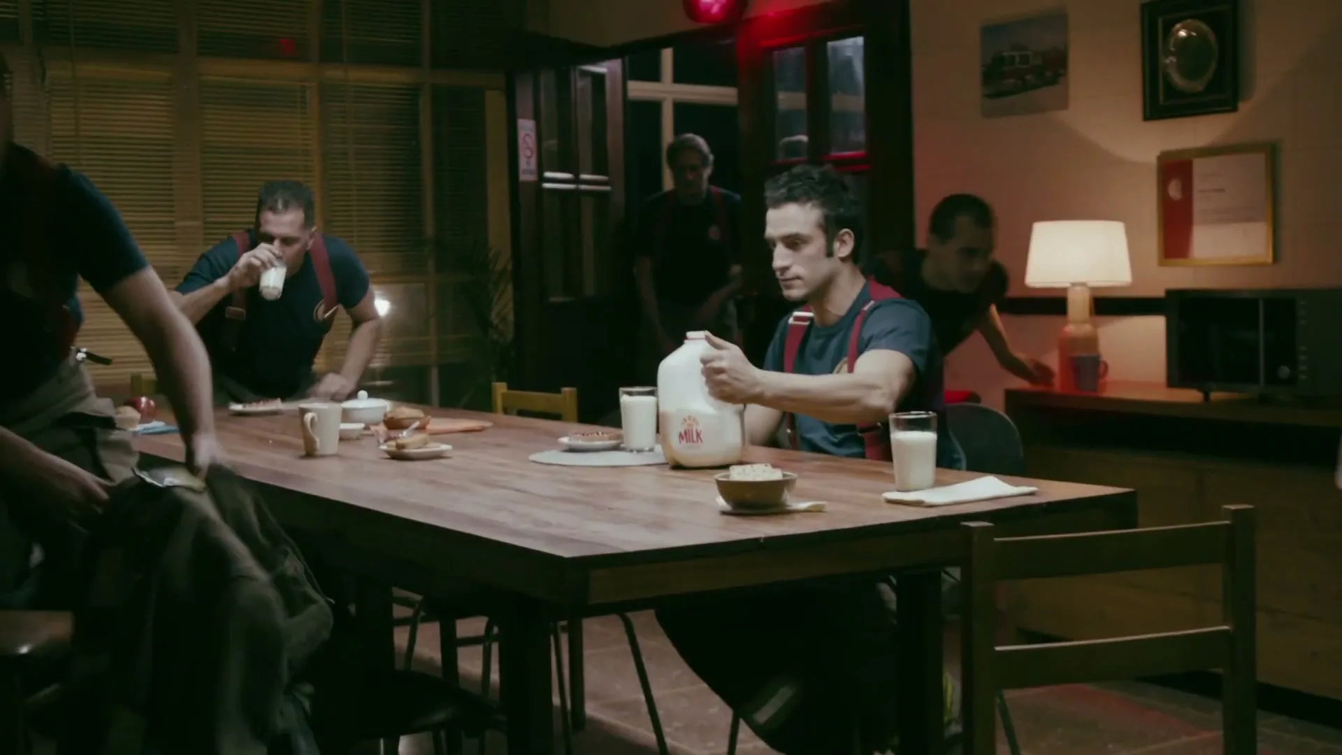 A group of male firefighters in uniform are sitting around a wooden table, eating and drinking milk, when an alarm sounds and they quickly get up to leave.