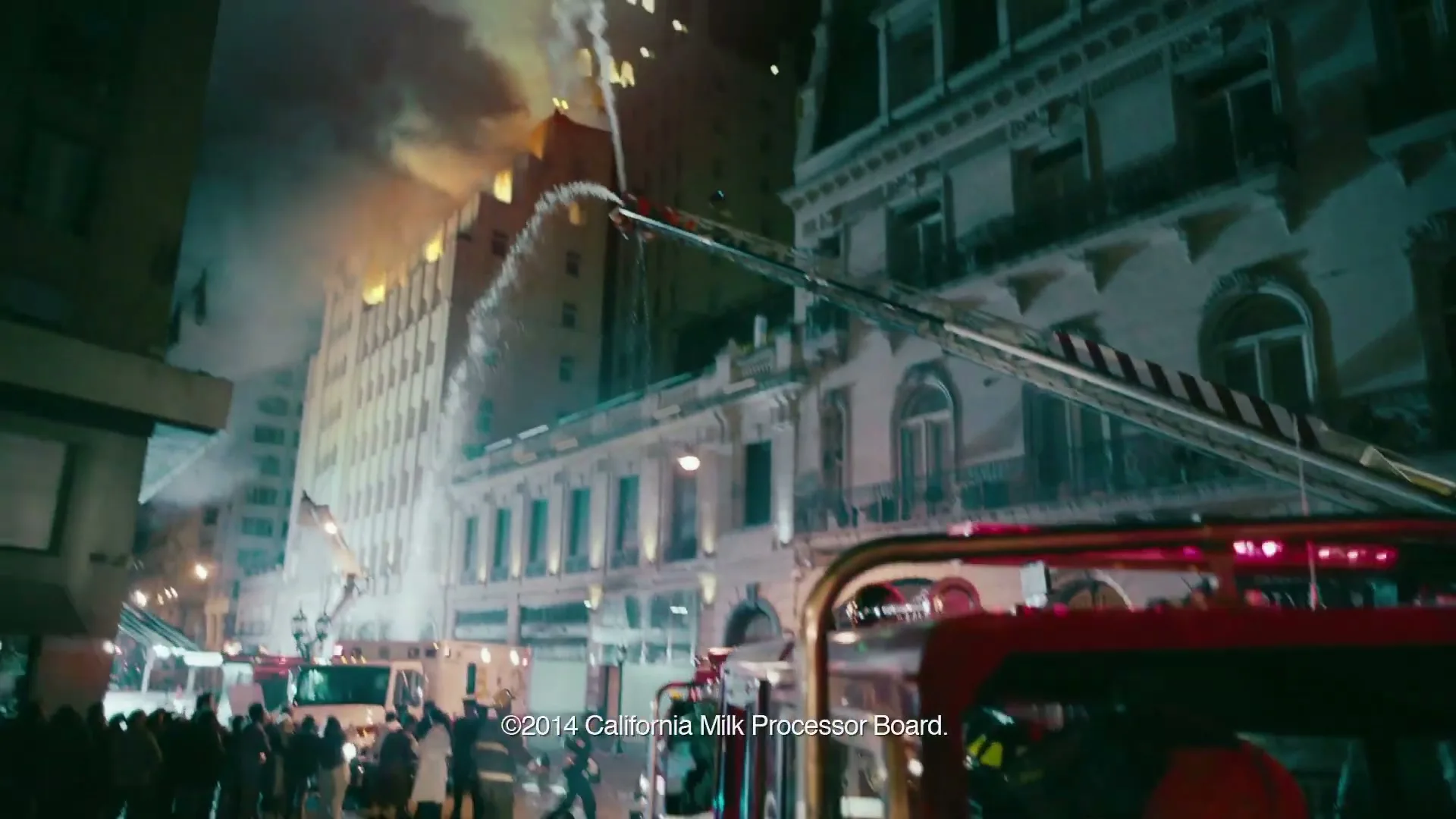 A young male firefighter in a red helmet and uniform sits in a fire truck, looking up at a burning building being sprayed with water at night.