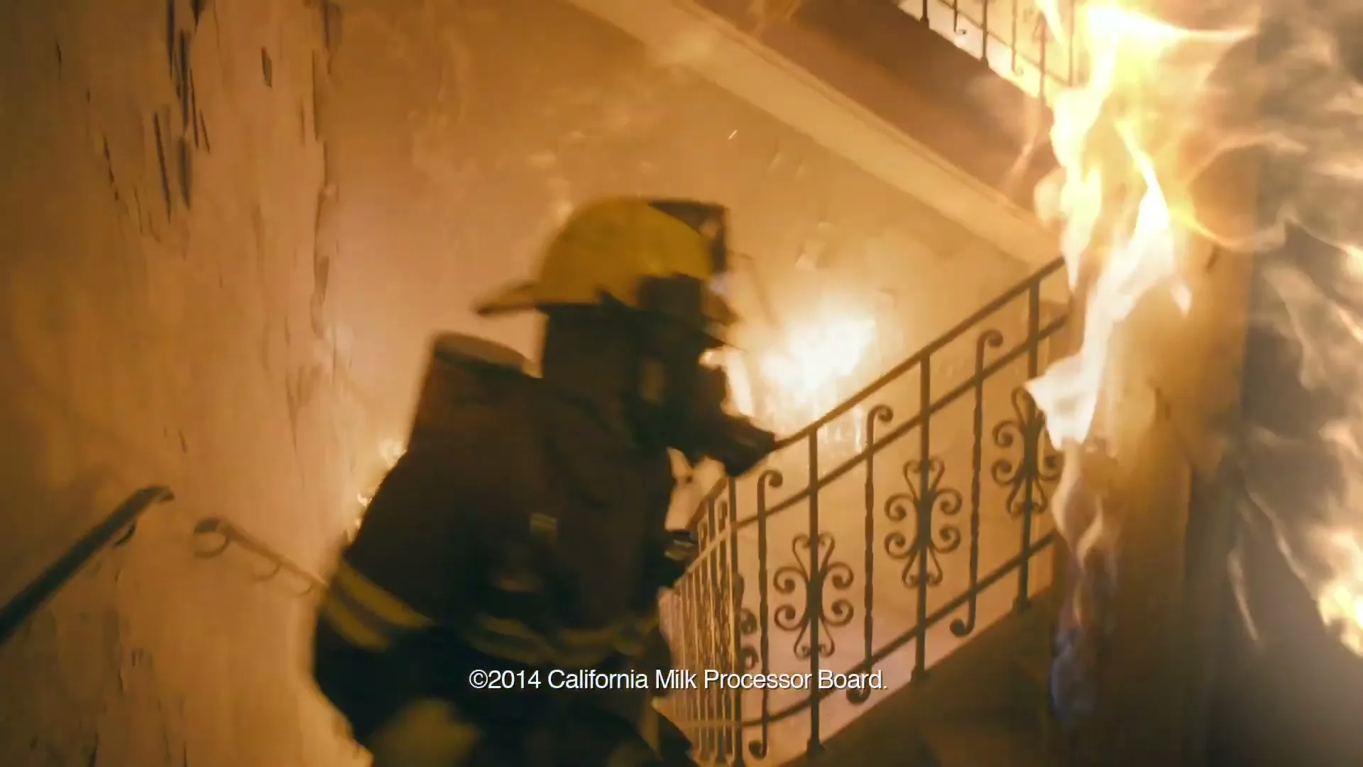 A firefighter in full gear runs up a burning staircase in a house, with flames engulfing the right side of the frame.