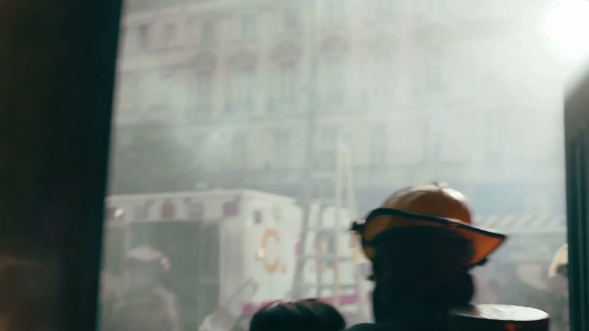 A firefighter in full gear, seen from behind, turns to face a burning building while other firefighters spray water in a smoky urban environment.