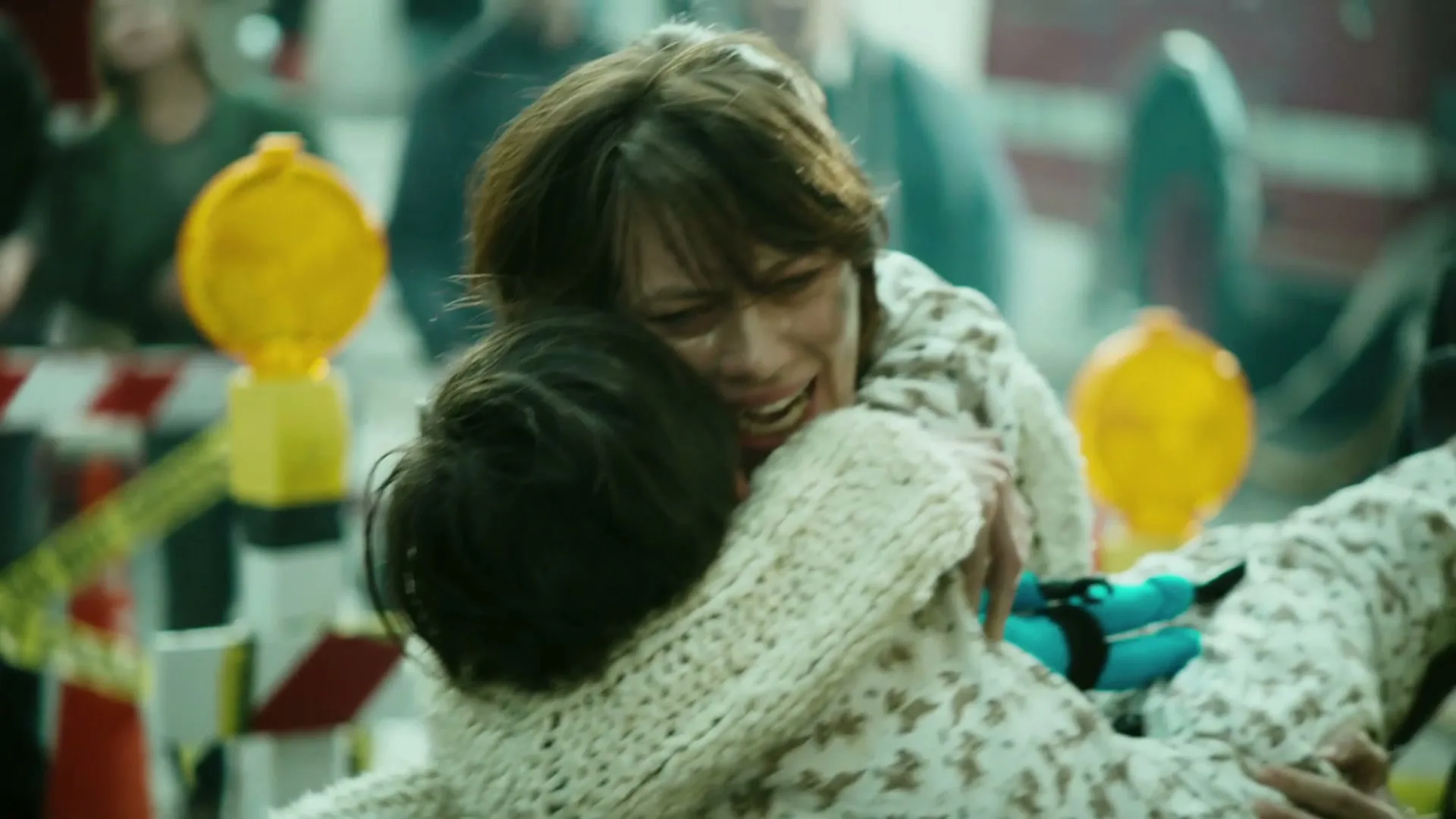 A woman with short brown hair, wearing a patterned top and knitted cardigan, cries while hugging a child in a chaotic, debris-filled urban street.