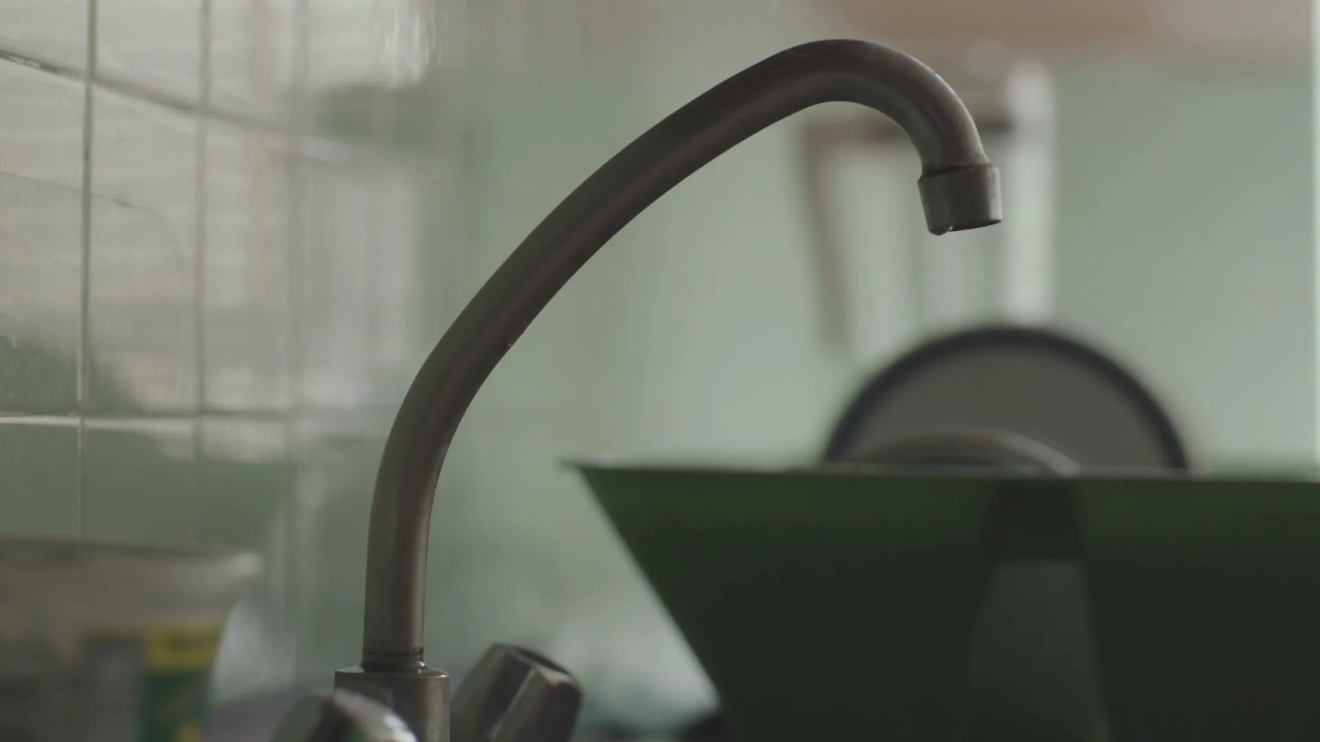 A close-up shot of a kitchen faucet with a single drop of water falling, next to a green dish rack.