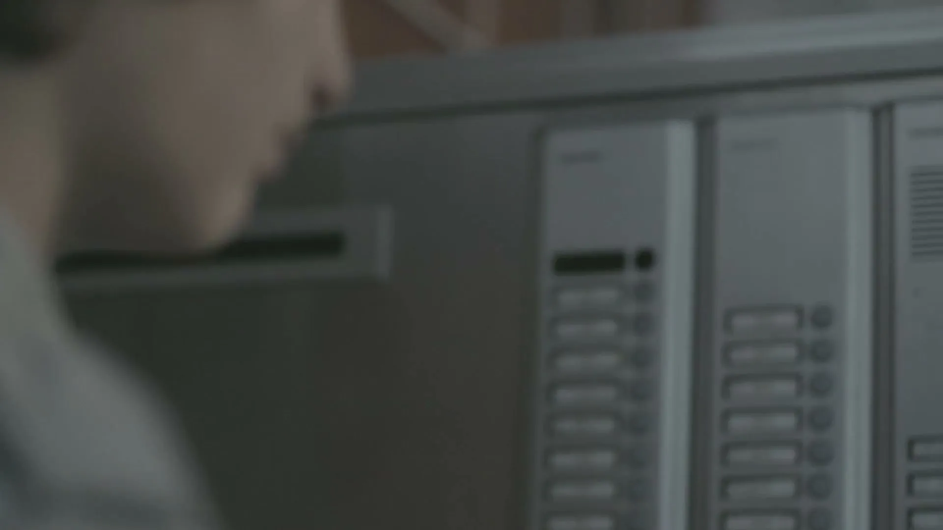 A young man with dark hair and a striped hoodie presses a button on a grey intercom panel in a building hallway.