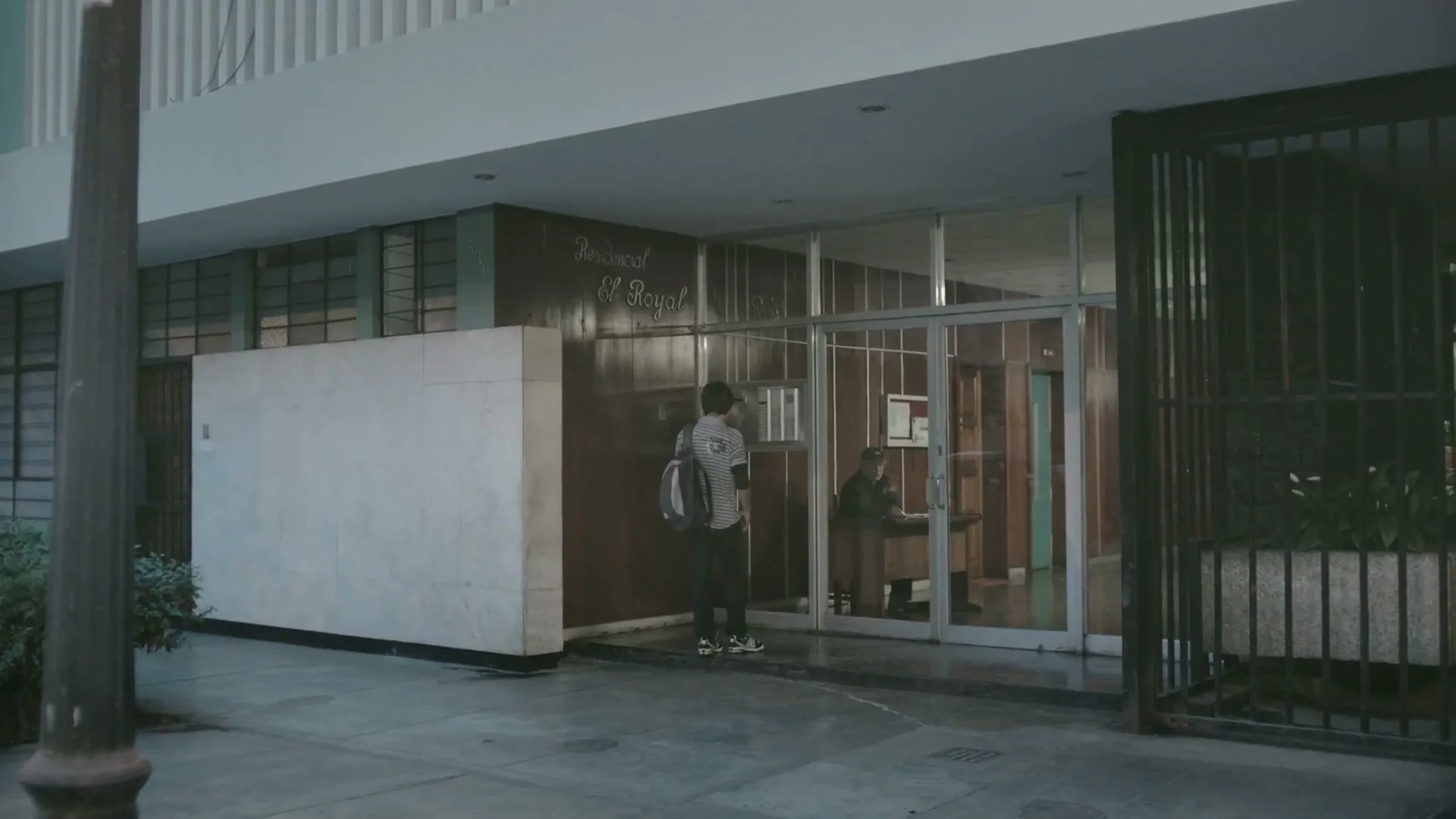 A young man with a backpack stands outside an apartment building entrance, looking inside where a woman sits at a desk.