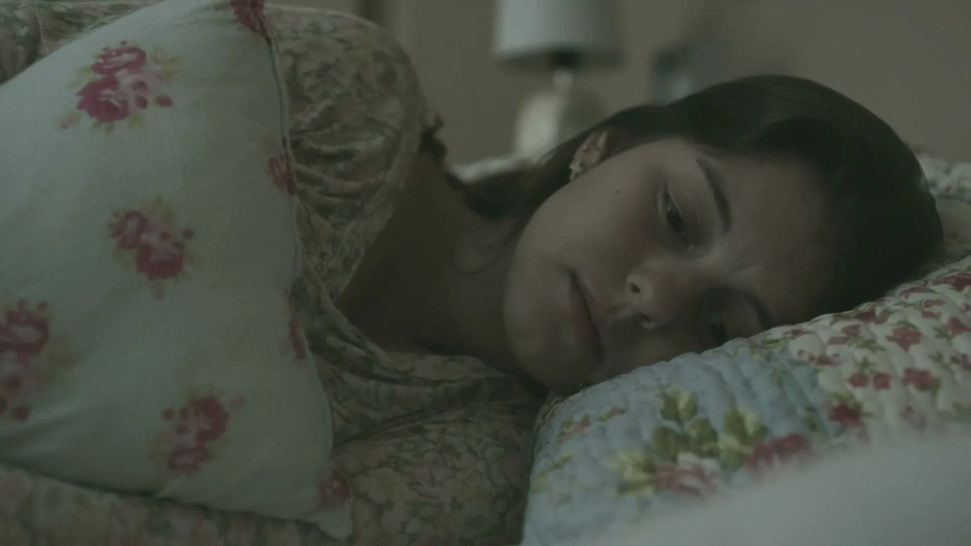 A young woman with dark hair sleeps soundly on a floral pillow in a dimly lit bedroom.