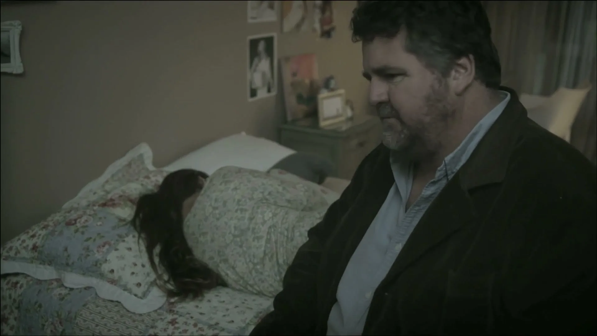 A middle-aged man with a beard sits on the edge of a bed, looking at a young woman lying face down, seemingly upset, in a bedroom.