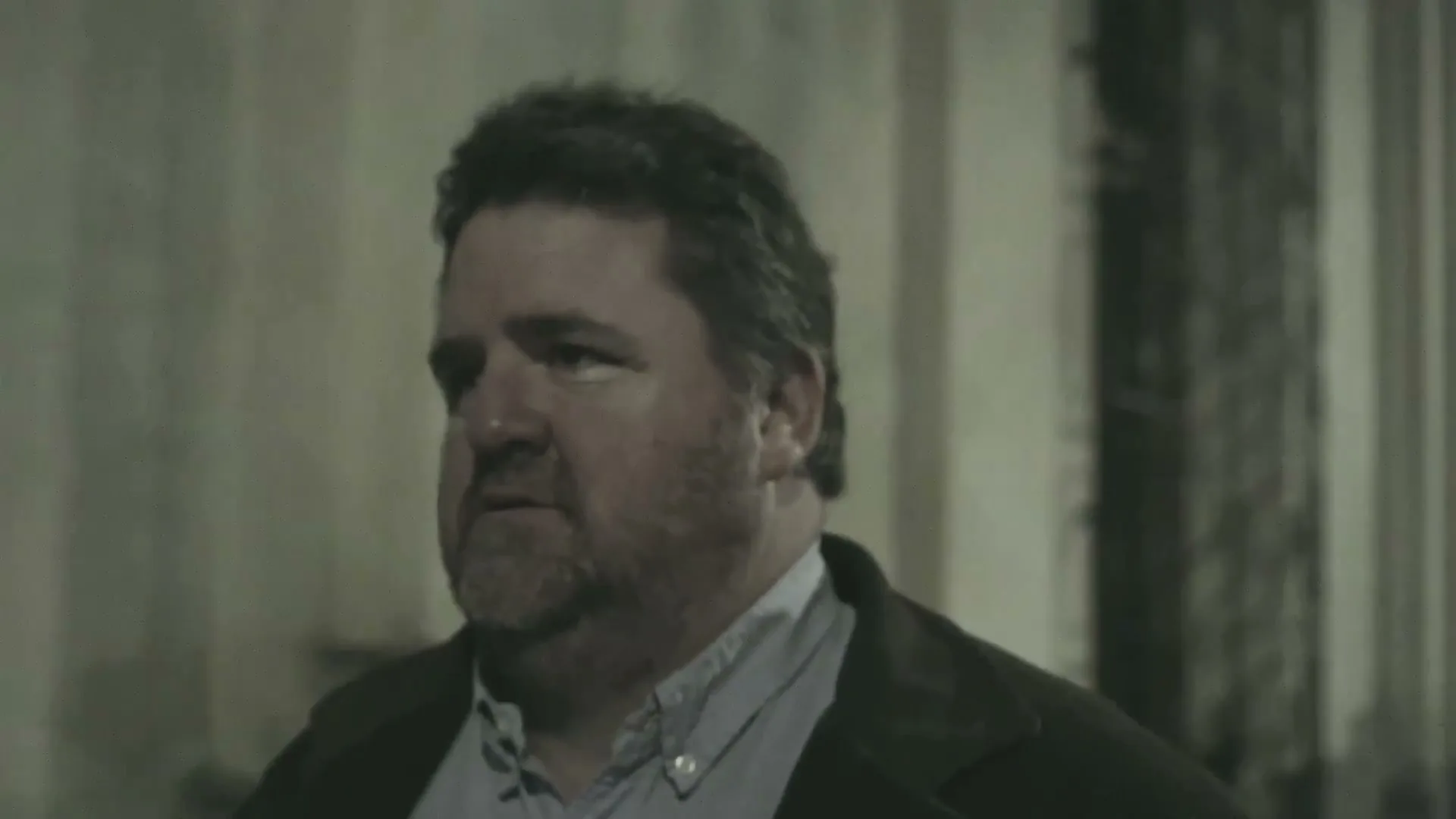 A middle-aged man with a beard and dark jacket walks through a dimly lit, urban exterior at night, looking around with a serious expression.