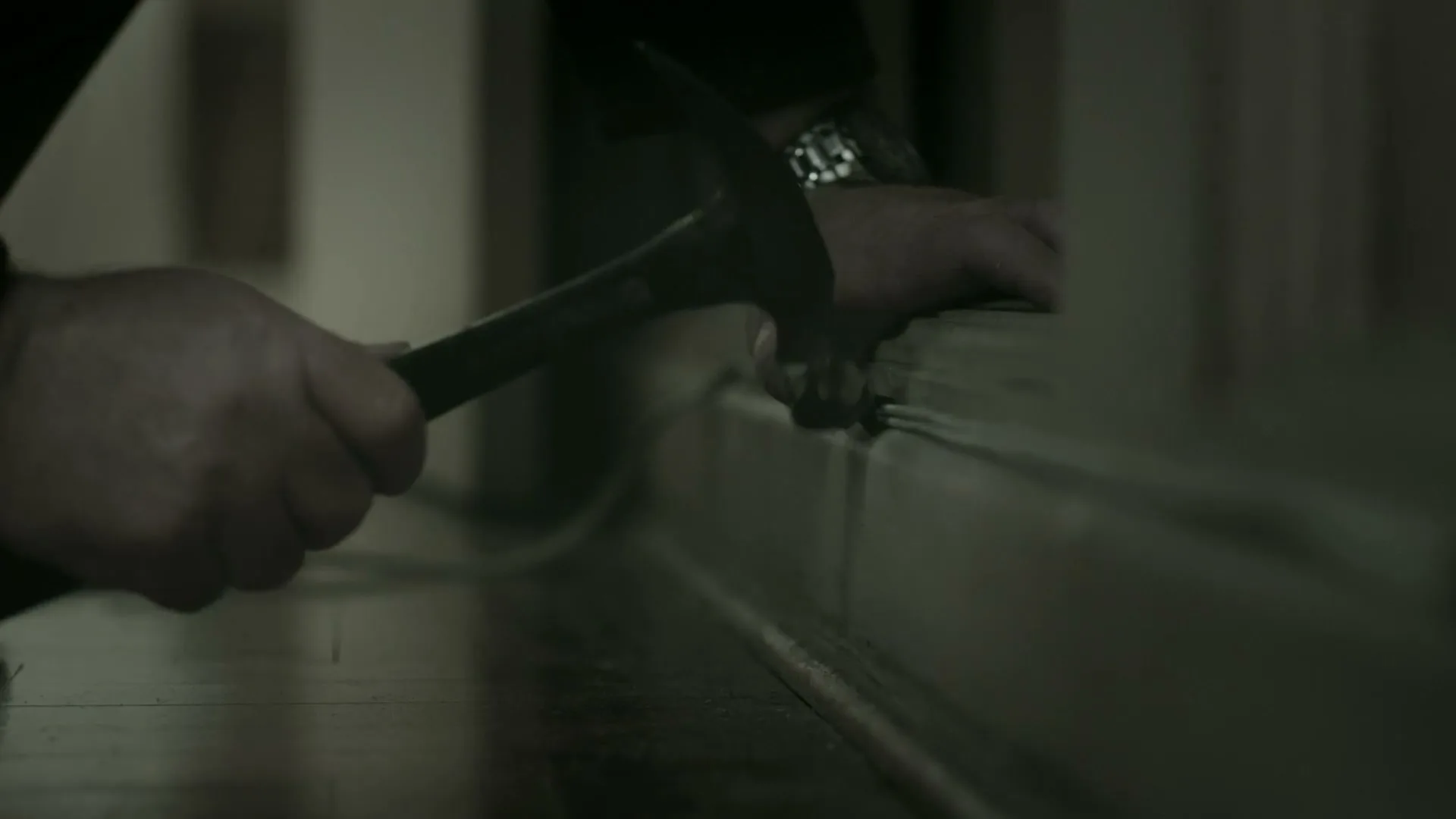 A man's hands are shown in a medium shot, using a hammer to secure a cable along a baseboard on a wooden floor in a dimly lit room.