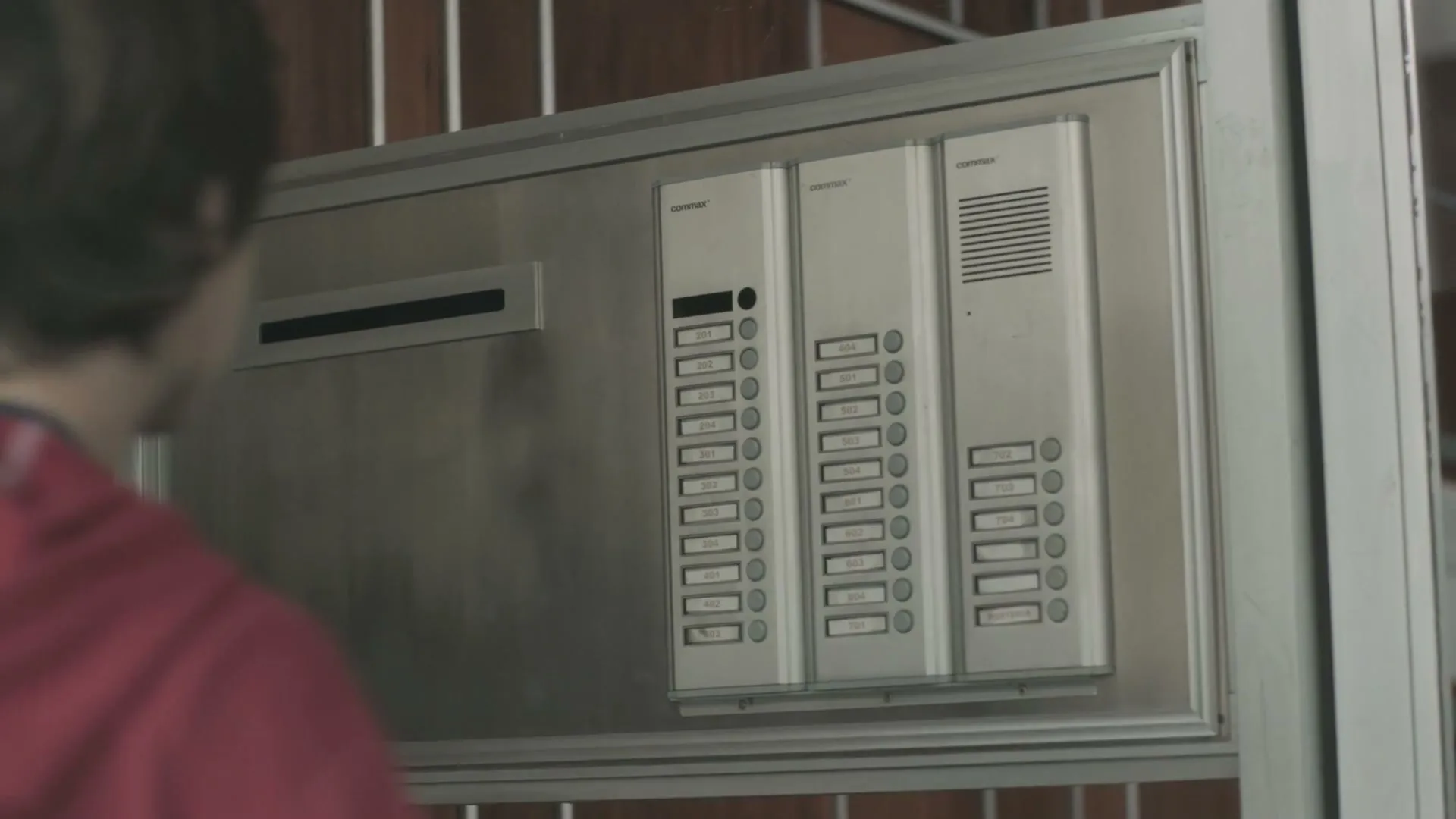 A young man in a red hoodie presses a button on a silver intercom panel outside an apartment building.
