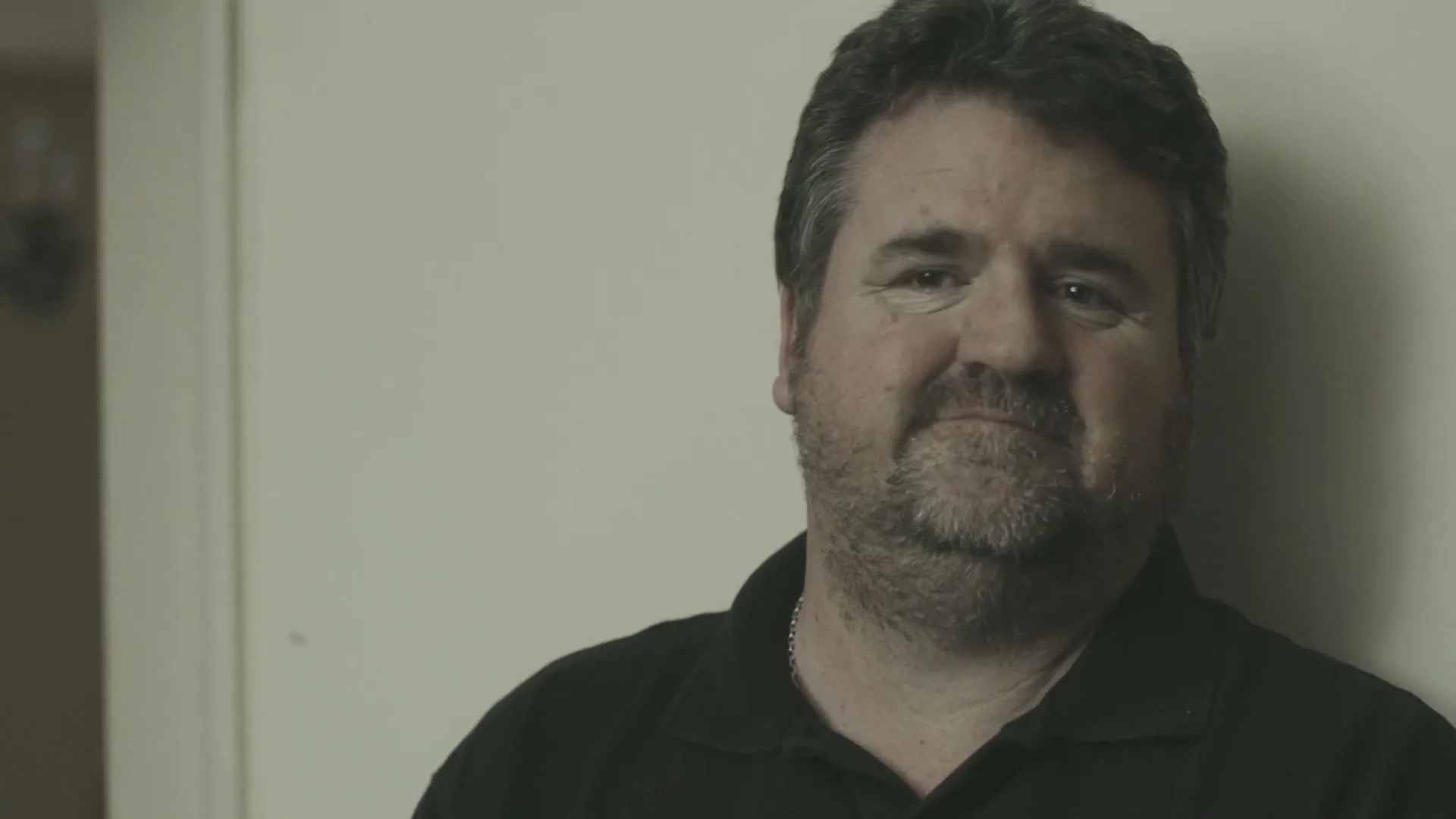 A middle-aged man with a beard, wearing a black polo shirt, leans against a wall with a slight smile on his face.