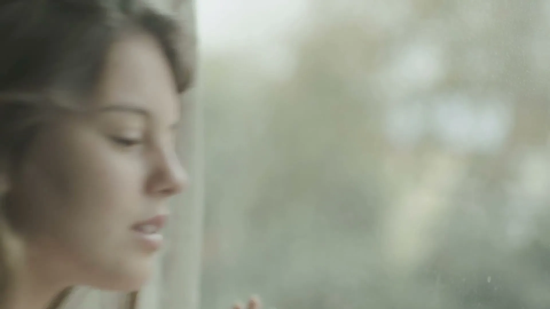 A young woman with long brown hair looks out a window, her face illuminated by soft natural light.