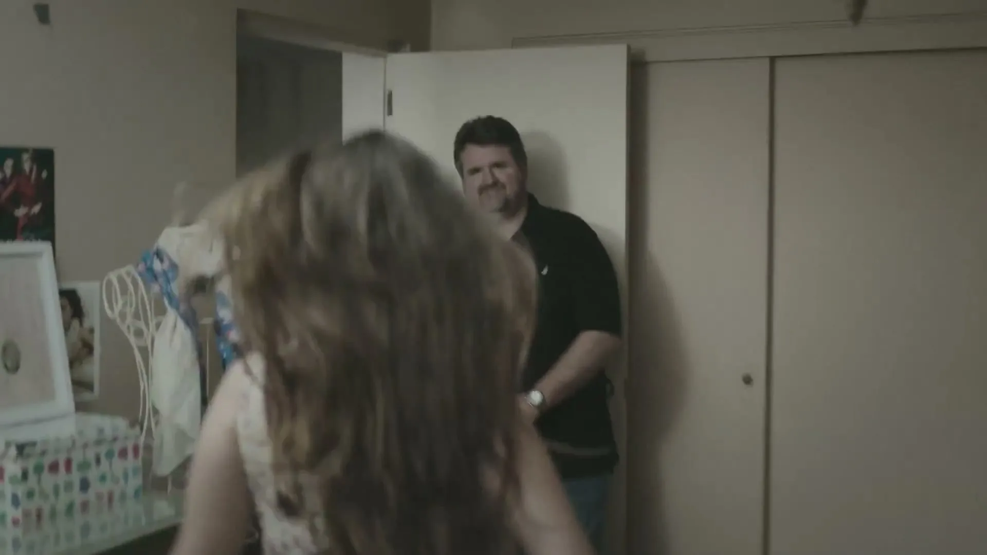A young woman with long brown hair turns and embraces a man in a black polo shirt, who is standing in a doorway in a bedroom.
