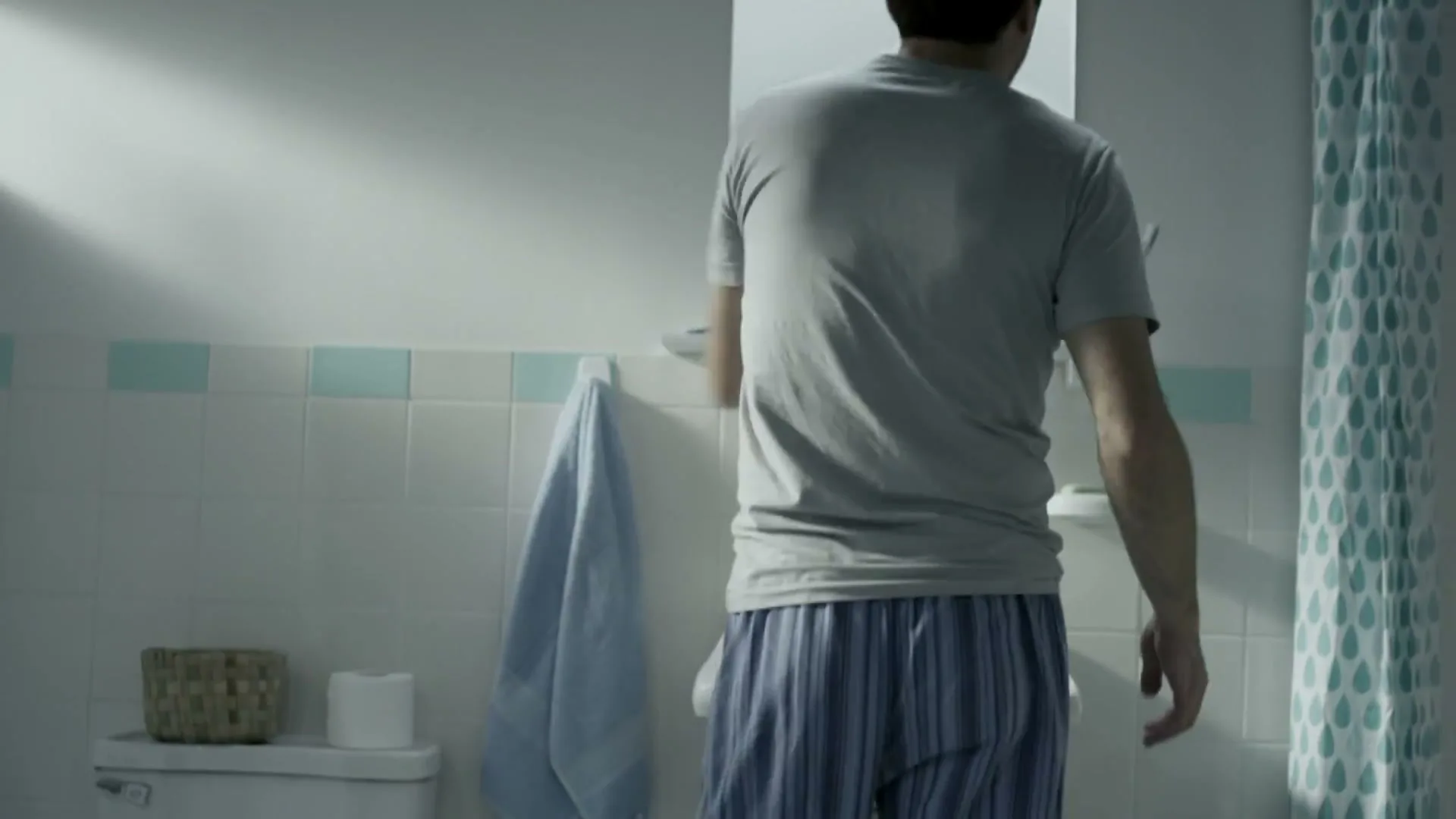 A man in a grey t-shirt and striped pajama pants stands with his back to the camera in a bathroom, reaching for something above a sink.