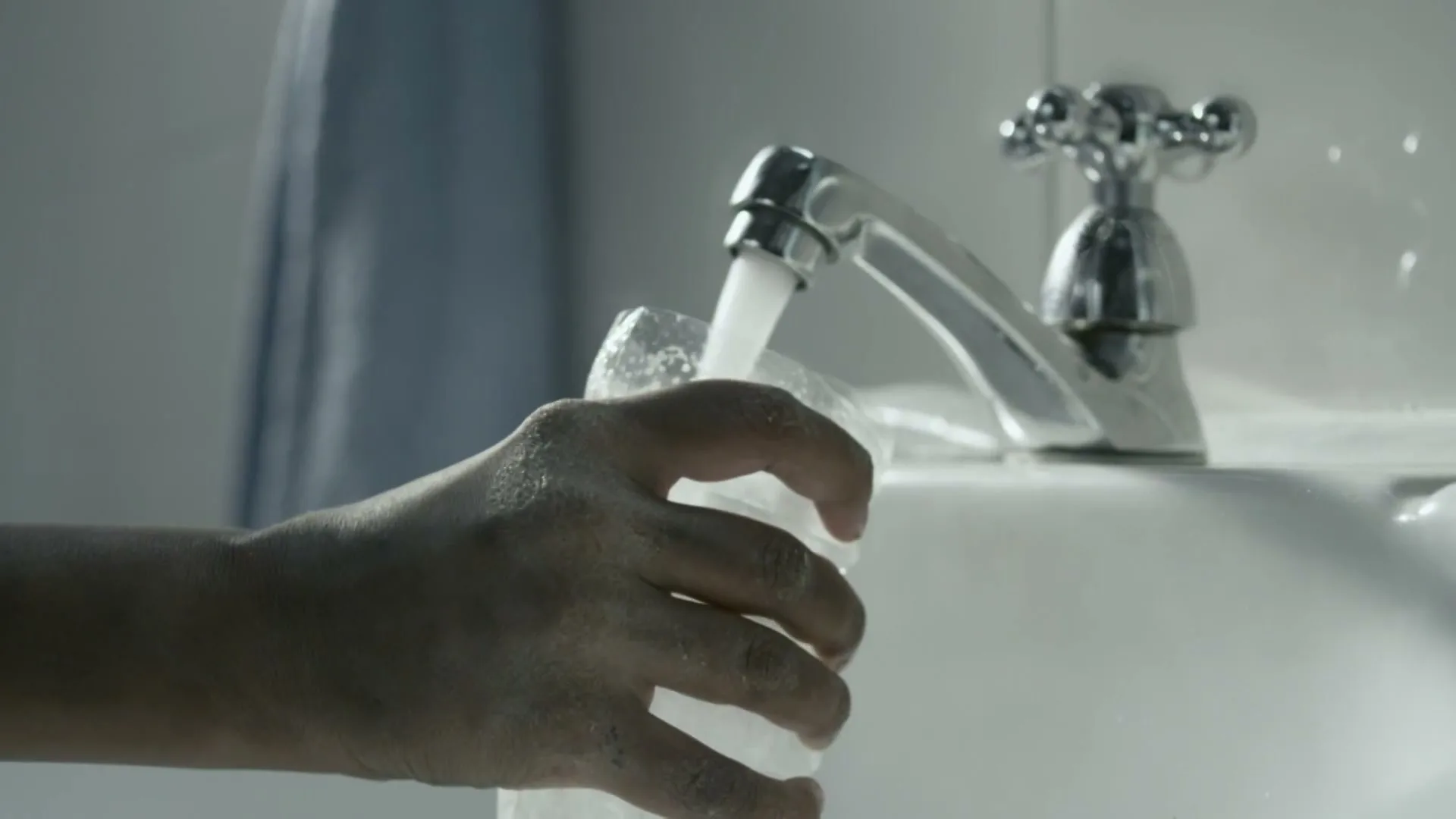 A person's hand holds a glass under a running faucet, filling it with water in a bathroom sink.