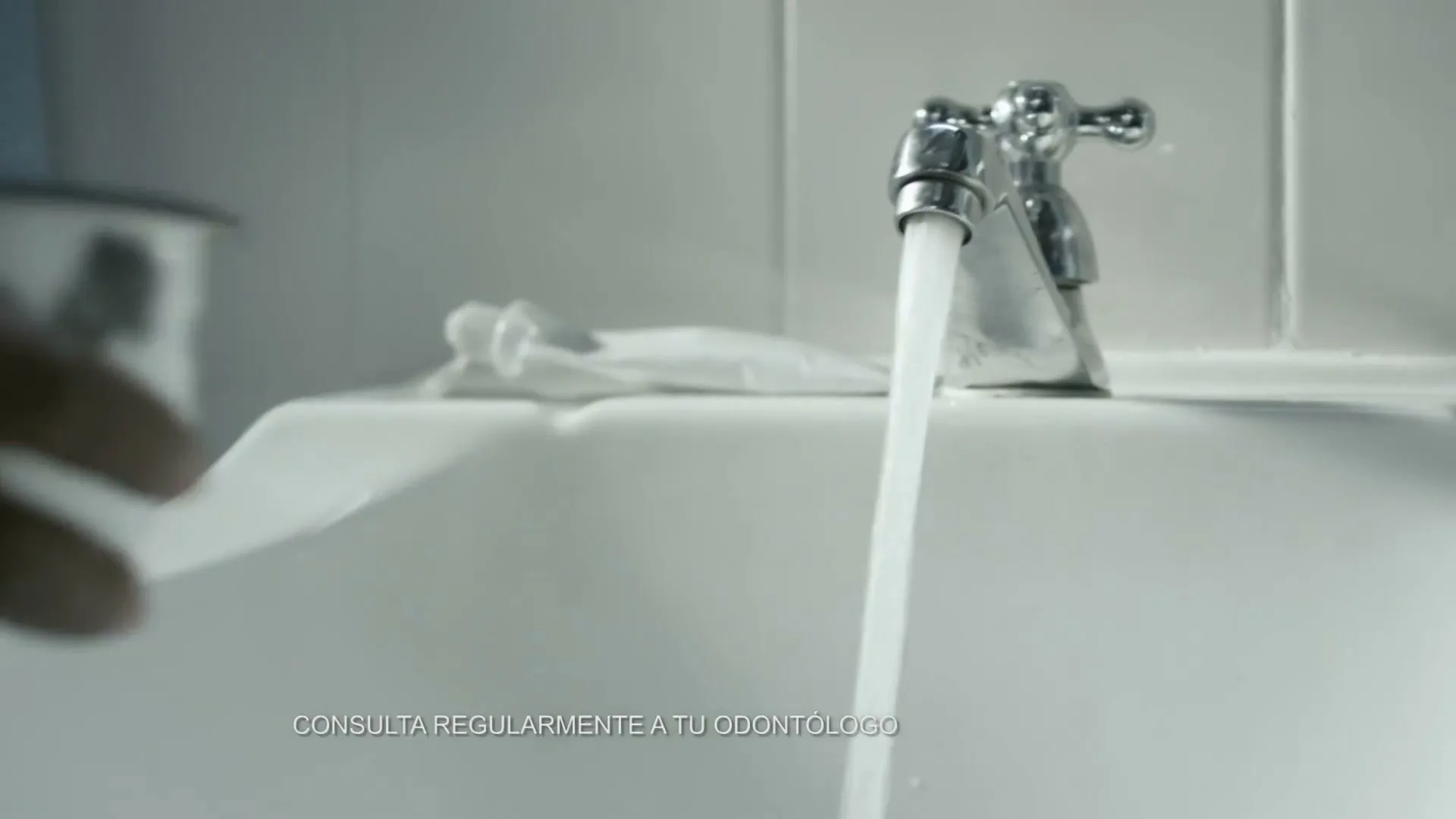 A person's hands are seen filling a small bowl with water from a running faucet in a bathroom sink, then washing a pear under the running water.