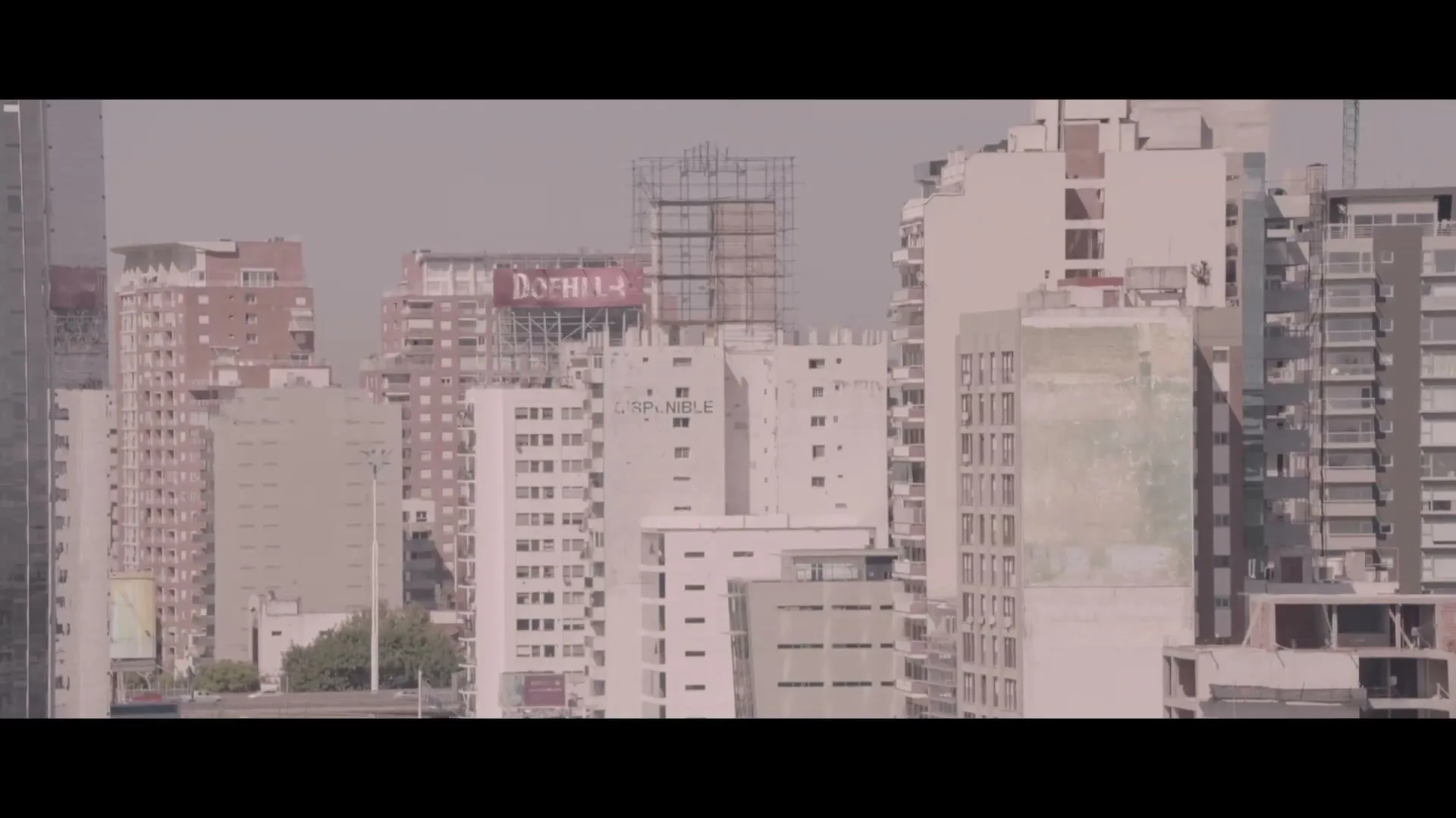 An establishing shot of a city skyline with numerous buildings and a billboard, under a clear sky during the day.