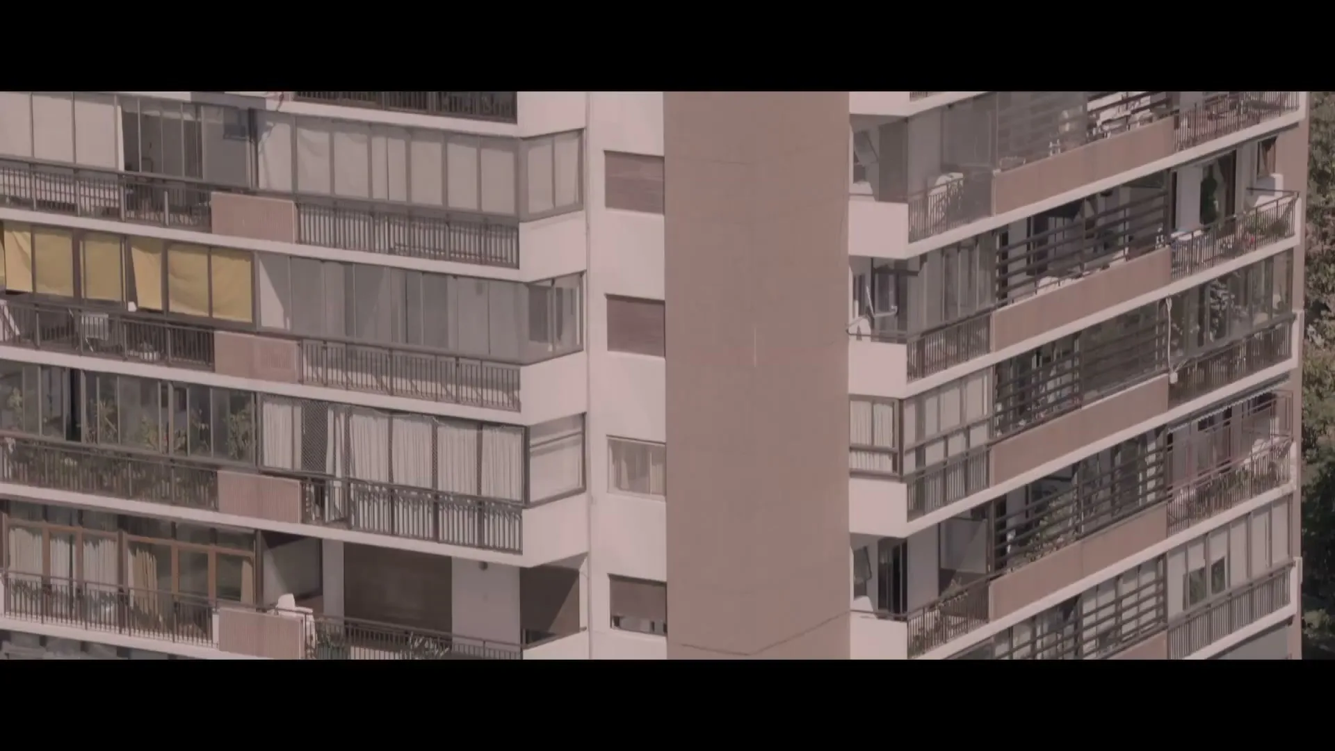 A high-angle shot captures the exterior of a tall residential building with multiple balconies and windows under a bright sky.