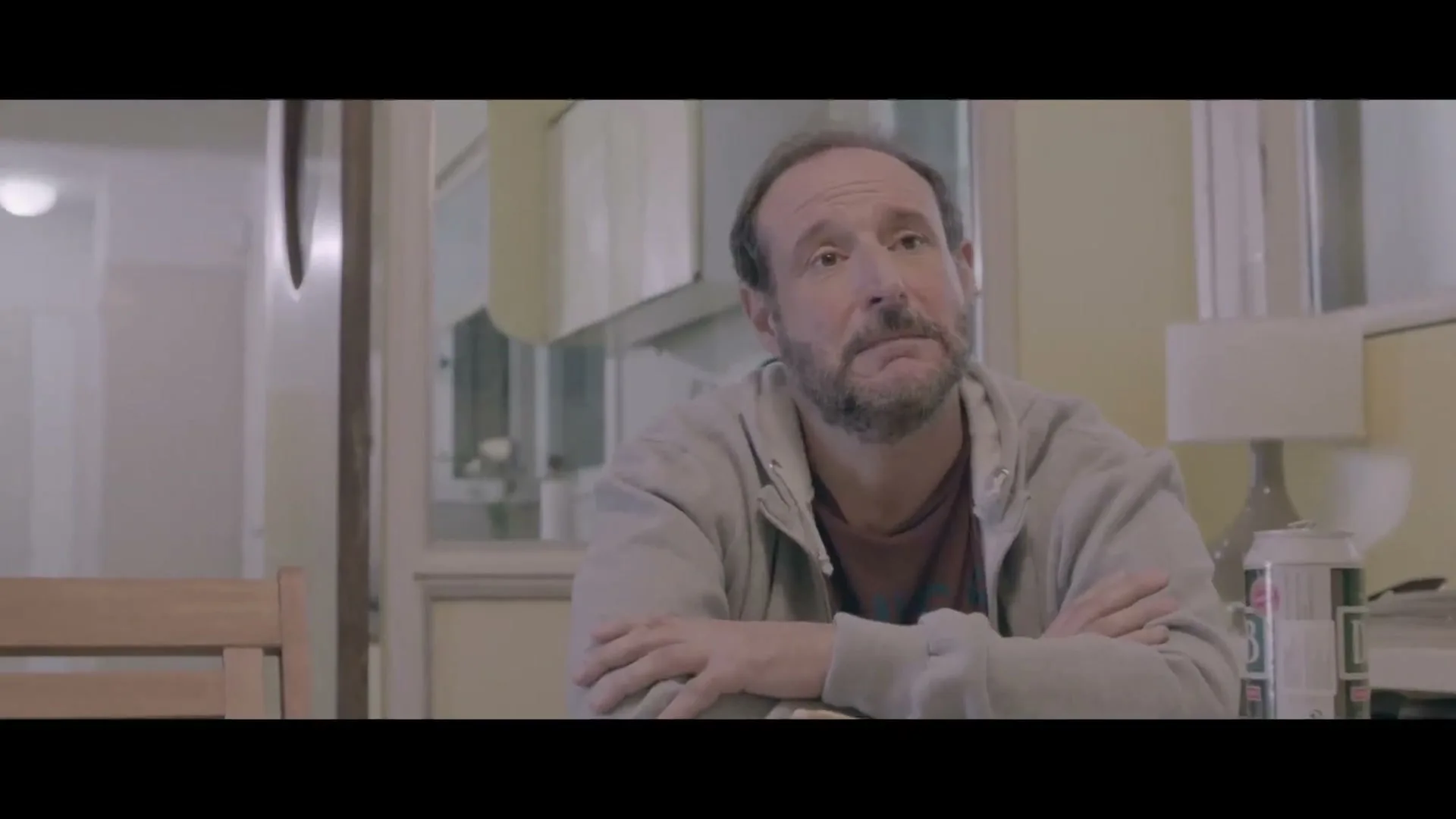 A middle-aged man with a beard sits at a kitchen table, looking up with a sad expression, while a young man walks by in the background.