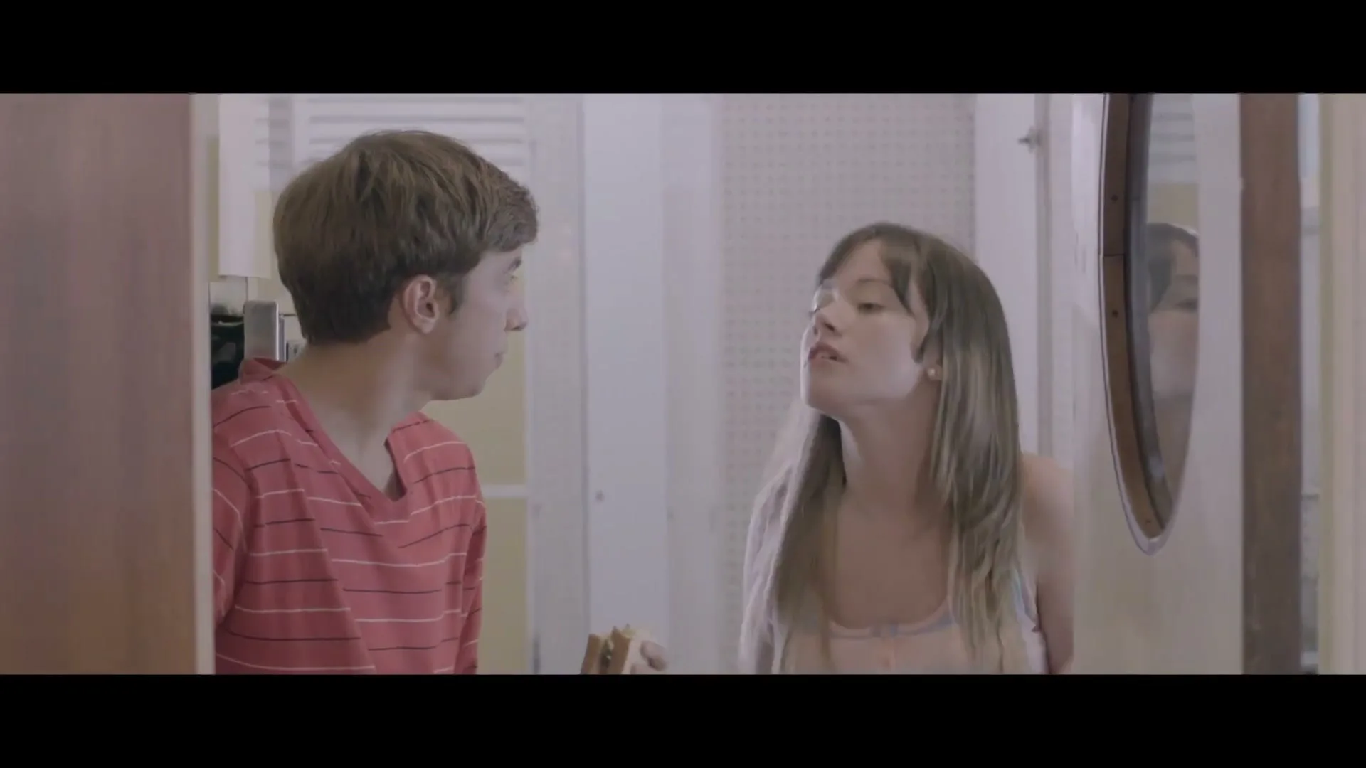 A young man in a red striped shirt eats a sandwich while talking to a young woman in a rainbow tank top in a hallway.