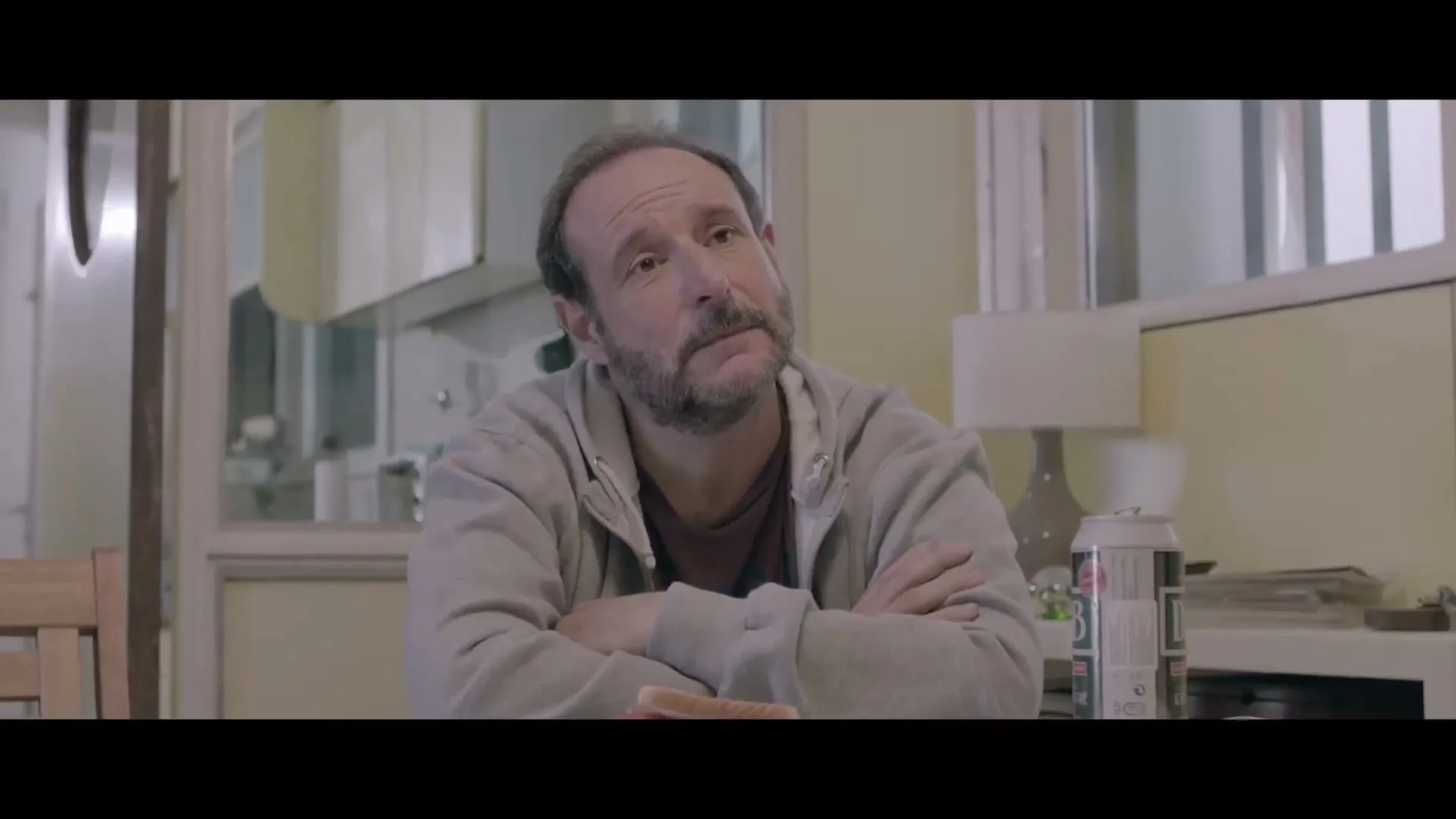 A middle-aged man with a beard sits at a table, looking up and to the right with a pensive expression in a softly lit room.