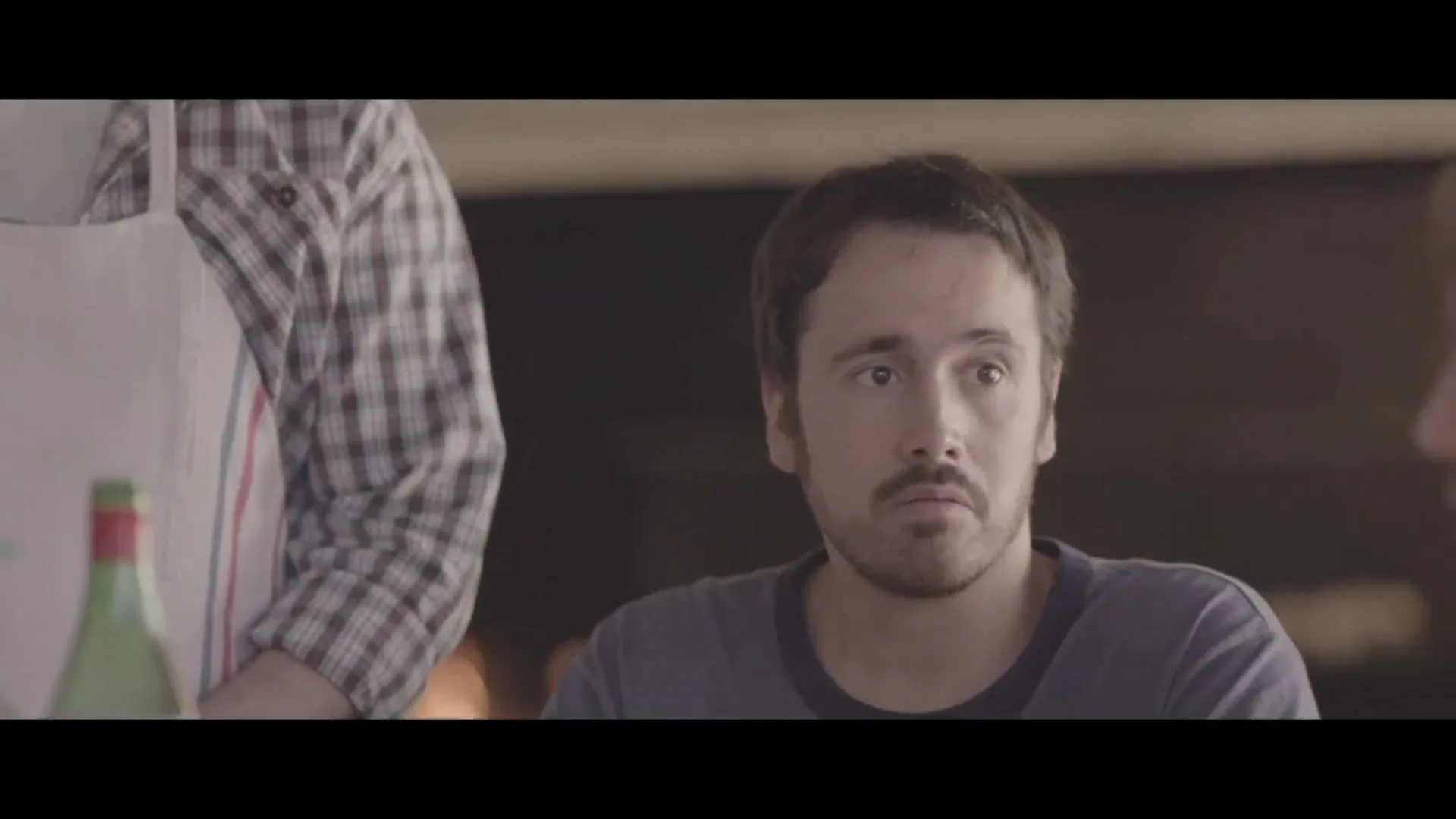 A man with dark hair and a beard looks surprised while sitting at a table, with another person in a plaid shirt and apron partially visible next to him.
