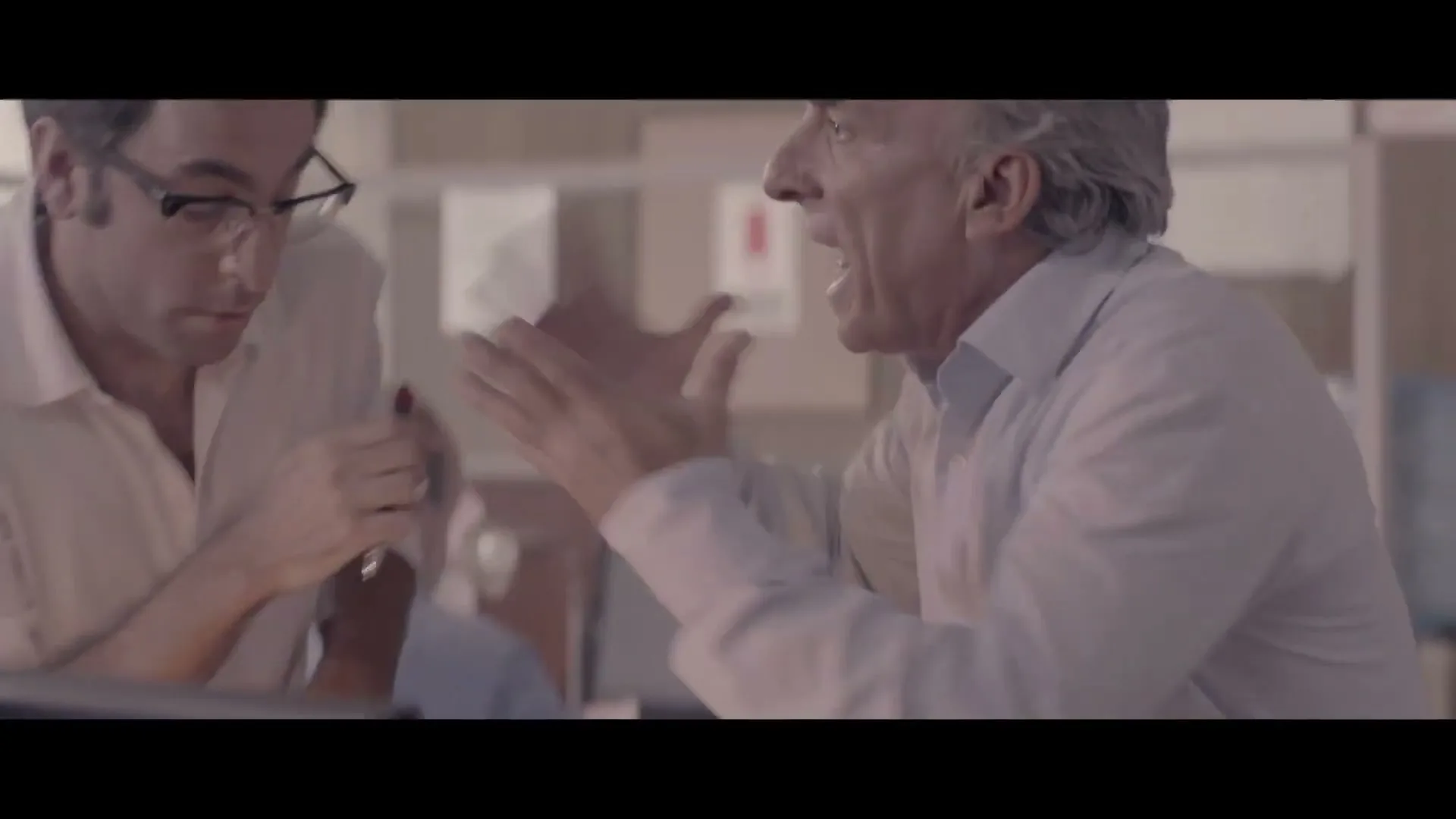 Two men in white shirts are working at a desk in an office, then one man gets angry and yells at the other, who looks annoyed.