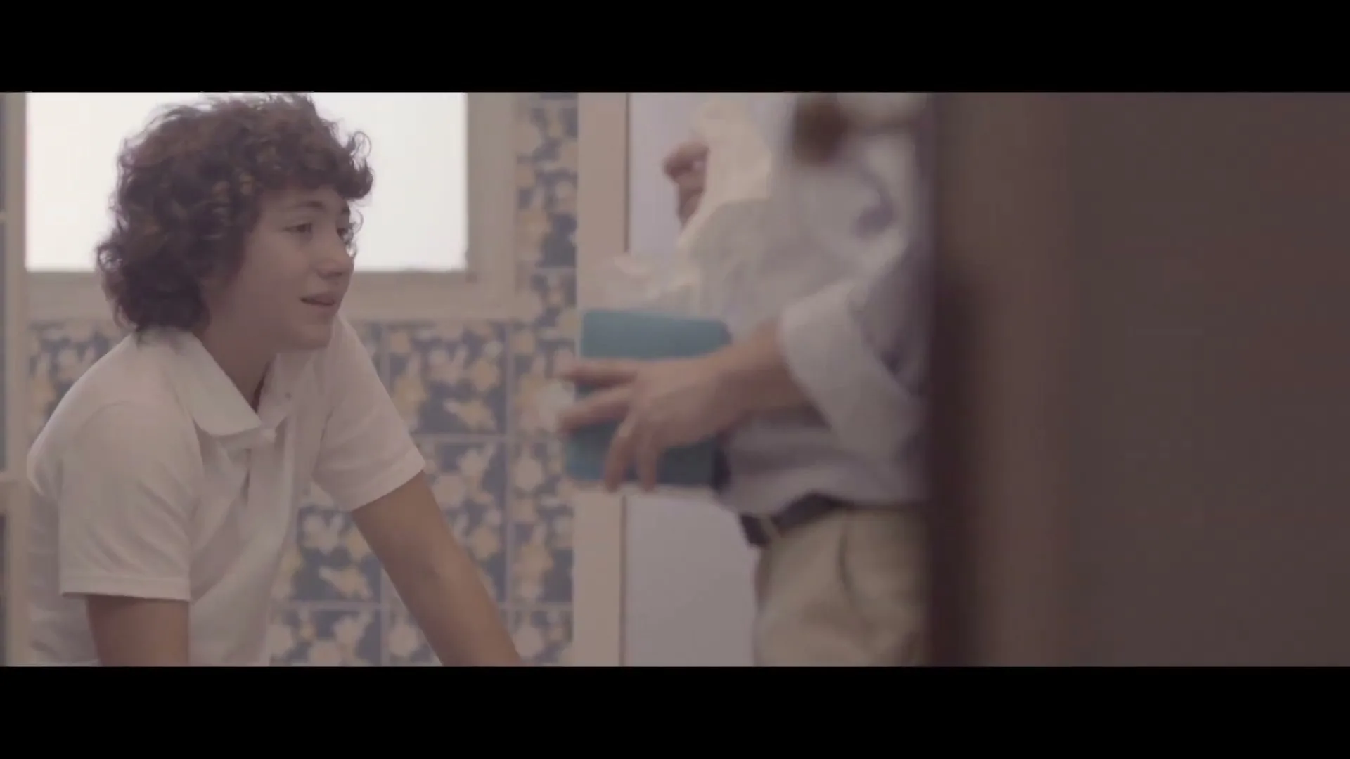 A young male with curly hair, wearing a white polo shirt, sits in a bathroom, looking sad and wiping his nose with a tissue, while an adult male offers him a box of tissues.