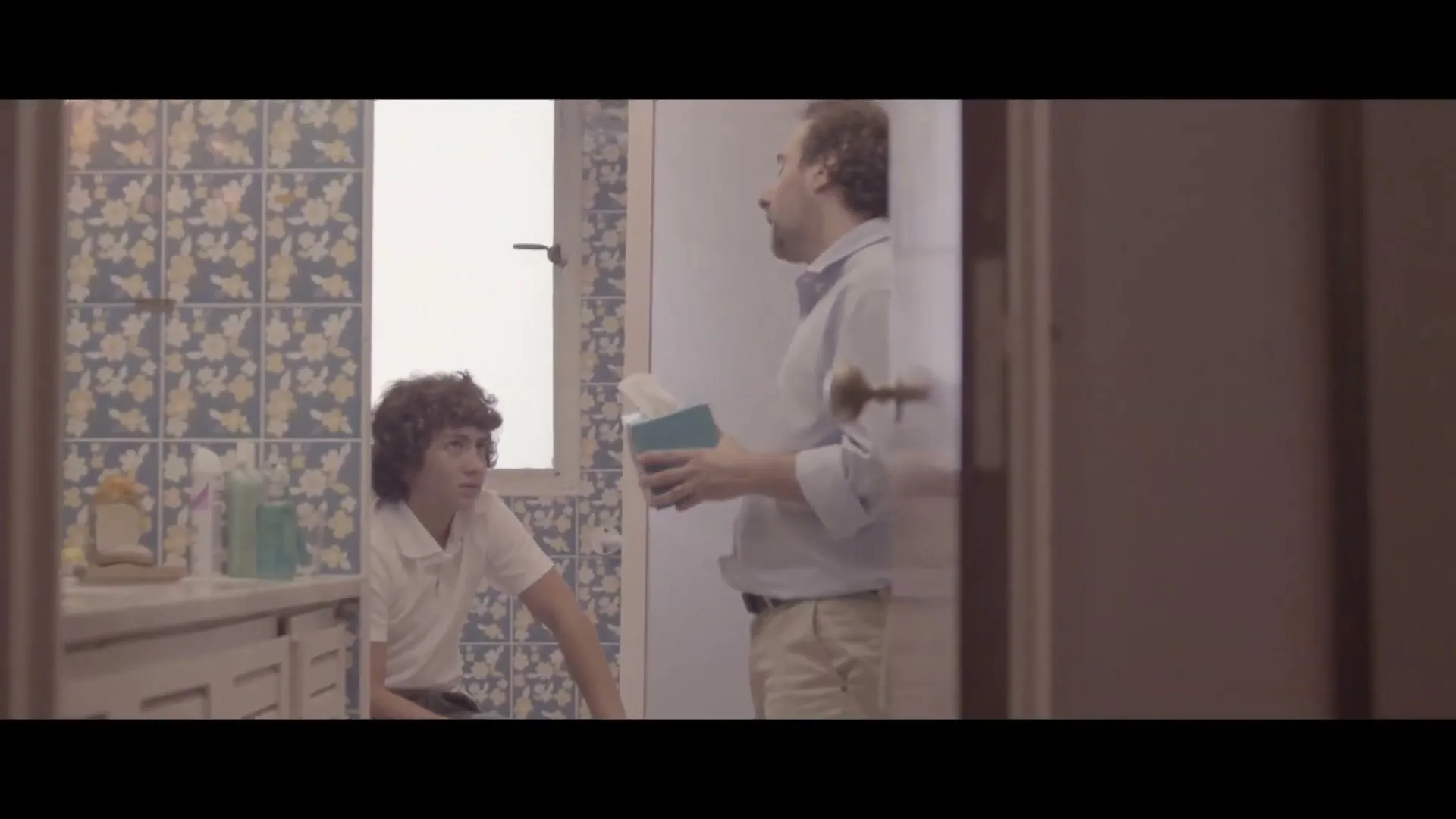 A father and son talk in a bathroom, the father holding a tissue box, the son sitting on the counter, seen through a doorway.