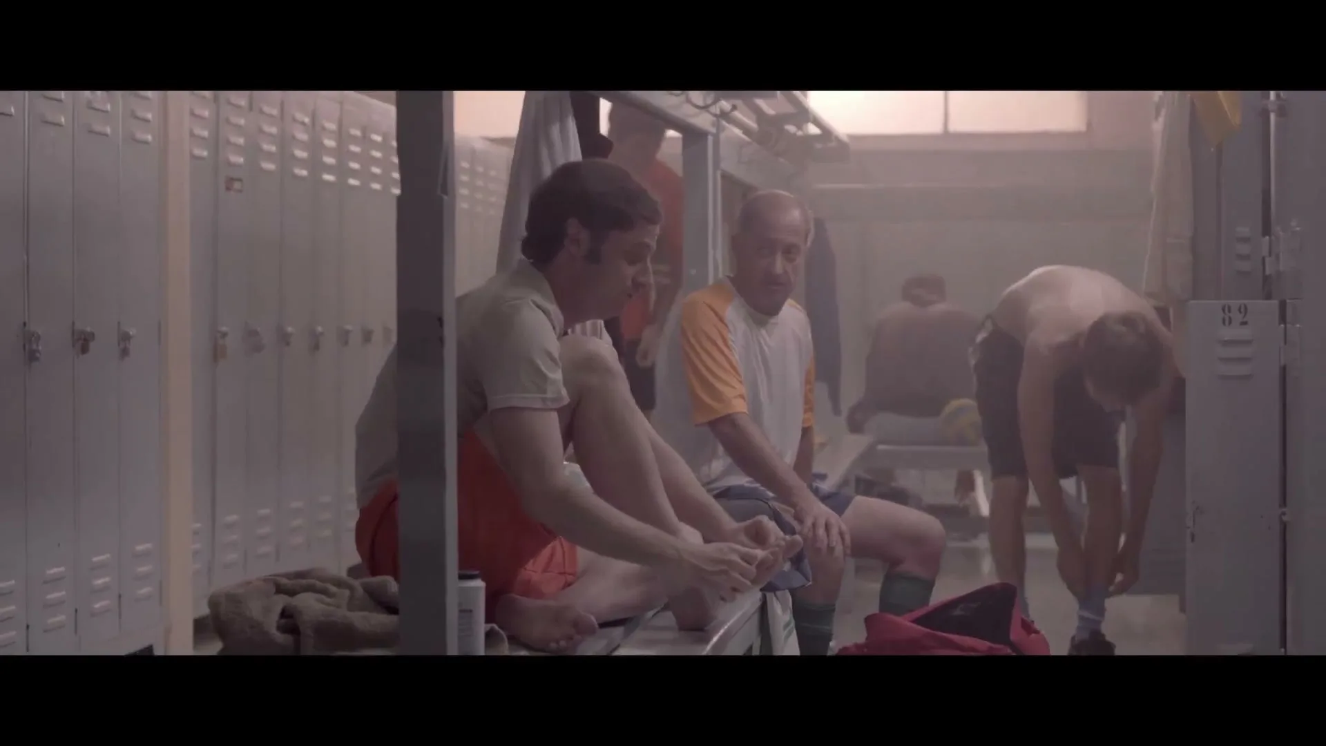 Two men sit on a bench in a locker room, putting on their socks and shoes, while other men are in the background.
