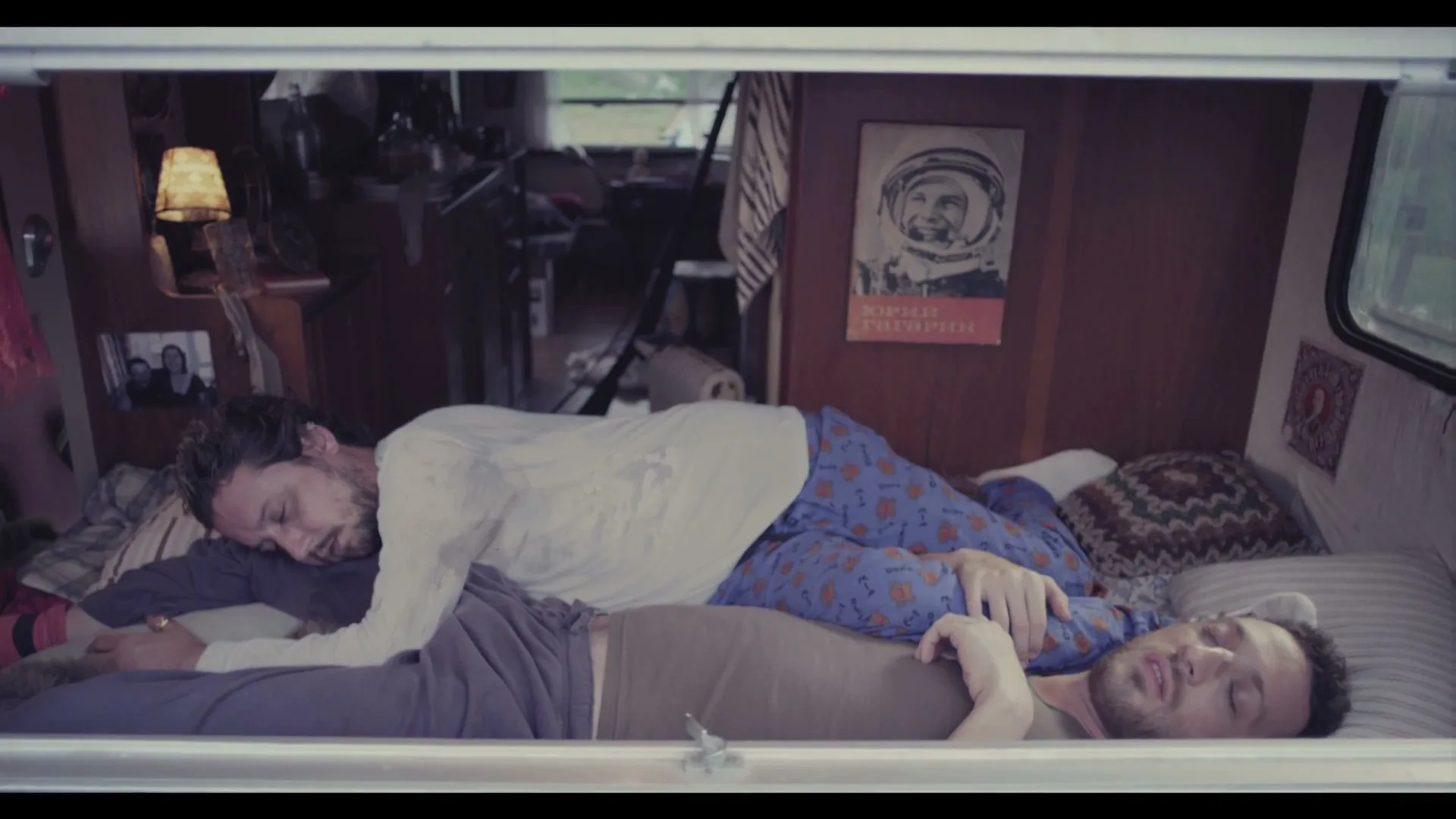 Two men sleep soundly in a bunk bed inside a camper van, one spooning the other, with a vintage space poster on the wall.