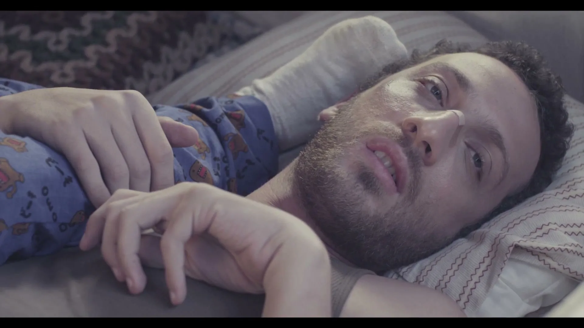 A man with a bandage on his nose and a beard lies in bed, wearing blue pajamas, looking tired and disoriented.