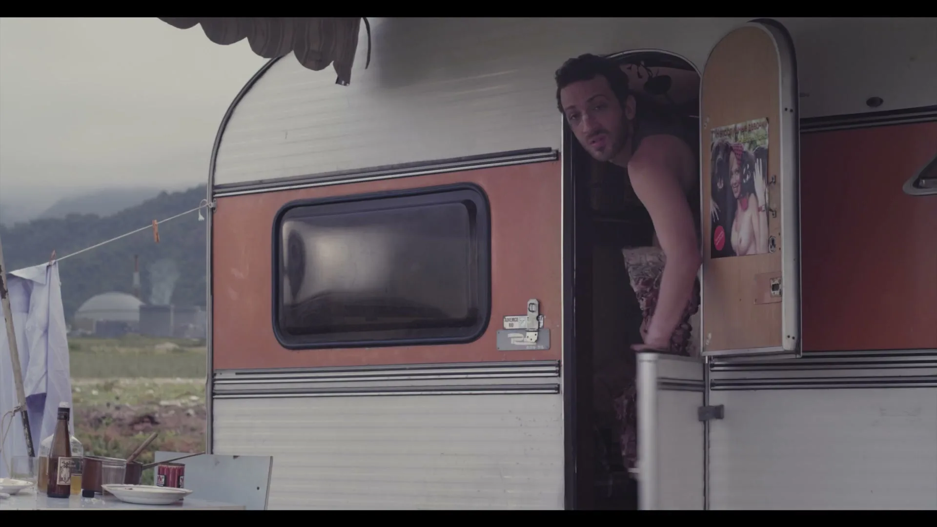 A man with dark hair and a beard, wearing a dark tank top, looks out of a camper door with a surprised expression, holding a large piece of raw meat.