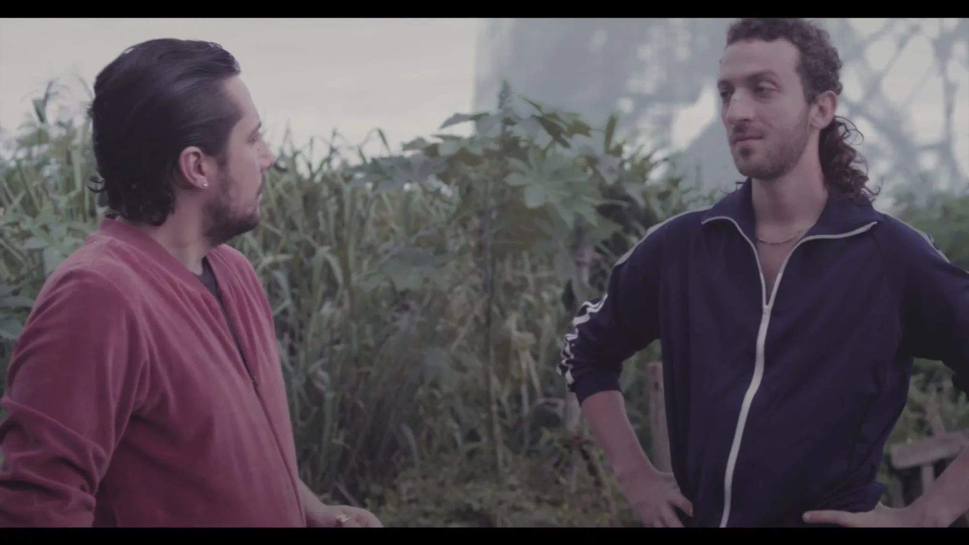 Two men, one in a red jacket and the other in a blue tracksuit, stand outdoors talking in a rural setting.