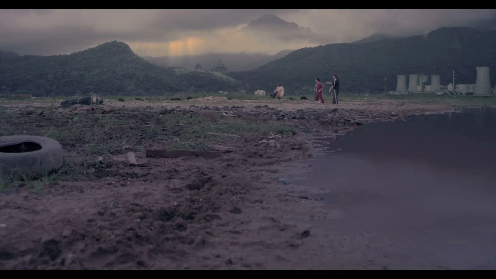 Two men walk along a muddy shore with a third man floating in the air behind them, against a backdrop of mountains and industrial buildings under a cloudy sky.