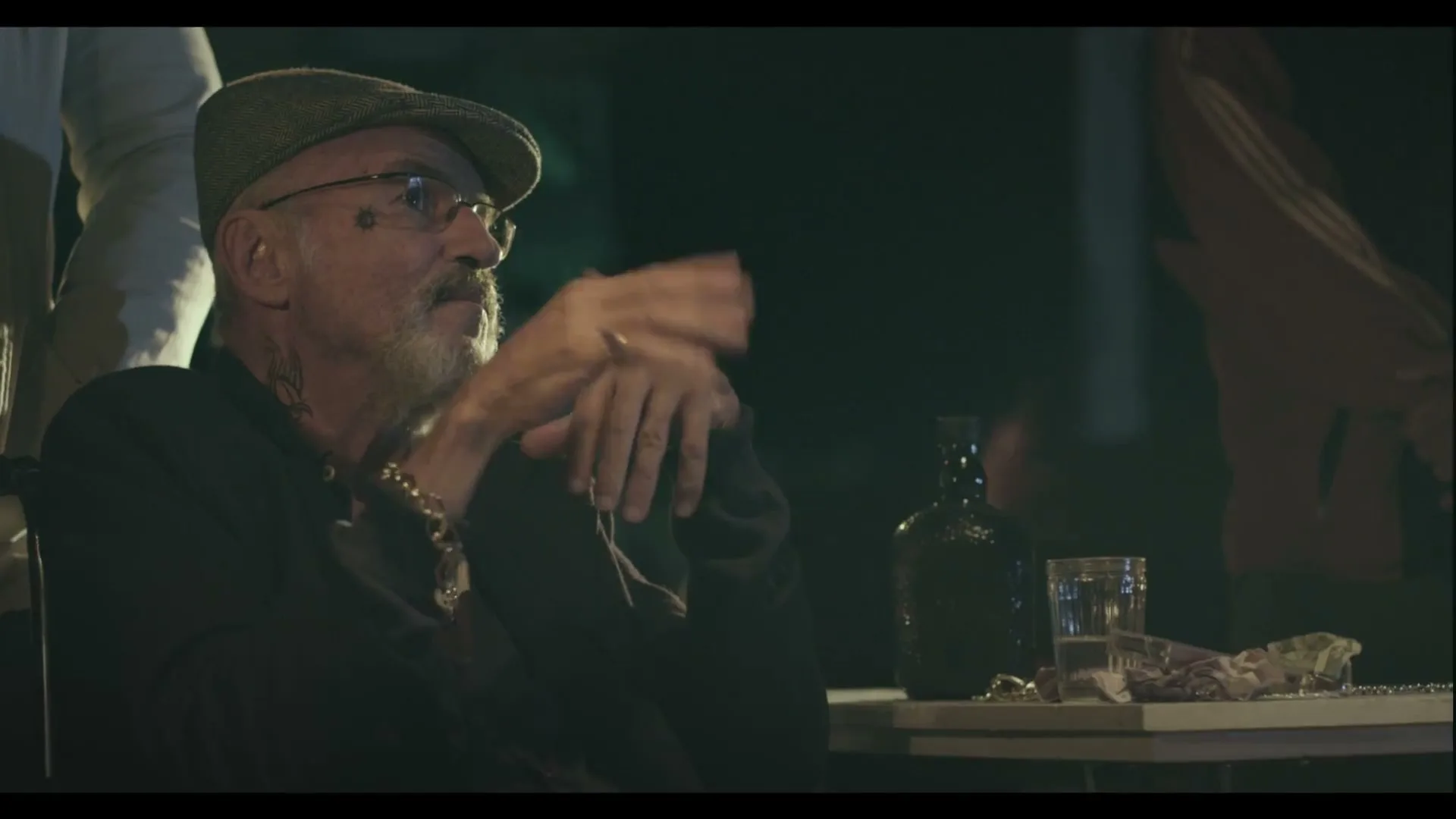 A man with a beard and glasses, wearing a flat cap, sits at a table with a bottle and glass, gesturing with his hands at night.