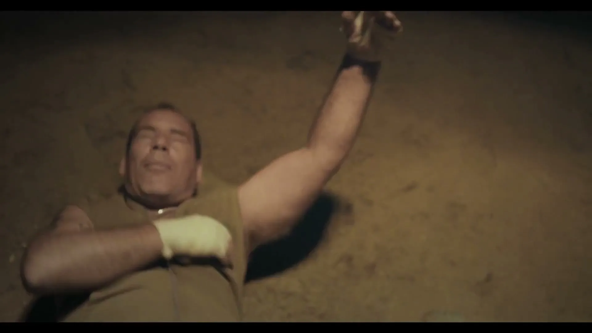 A male boxer in his 40s, wearing a sleeveless hoodie and hand wraps, collapses to the ground in a dimly lit room.