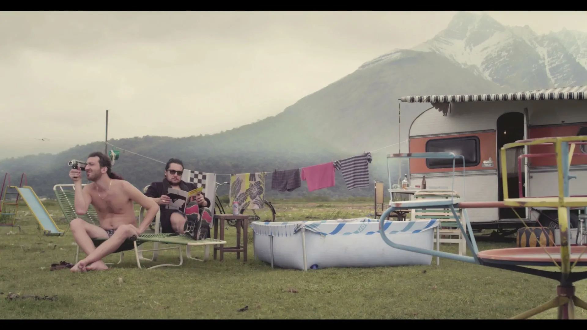 Two men relax outside a vintage camper van in a grassy field with mountains in the background; one takes photos, the other reads.