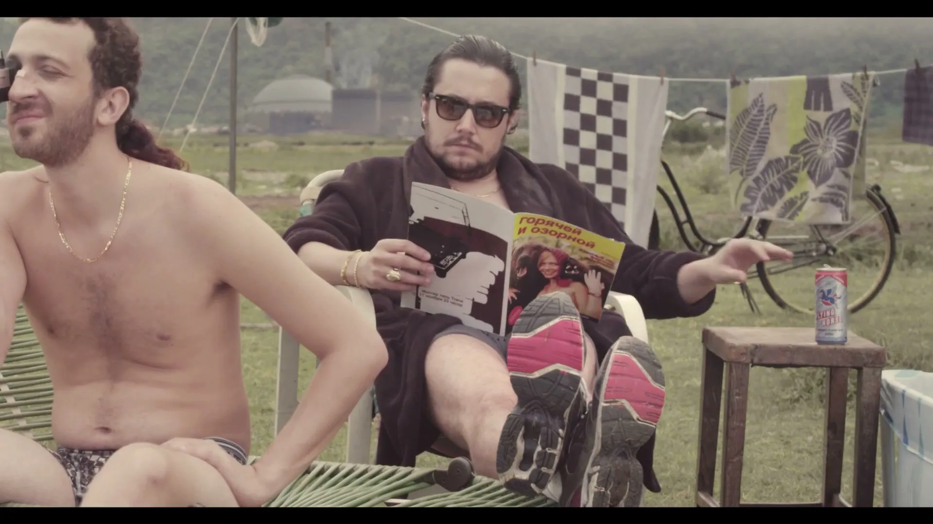 Two men relax outdoors; one reads a magazine while the other sits shirtless, both enjoying a leisurely moment in a rural setting.