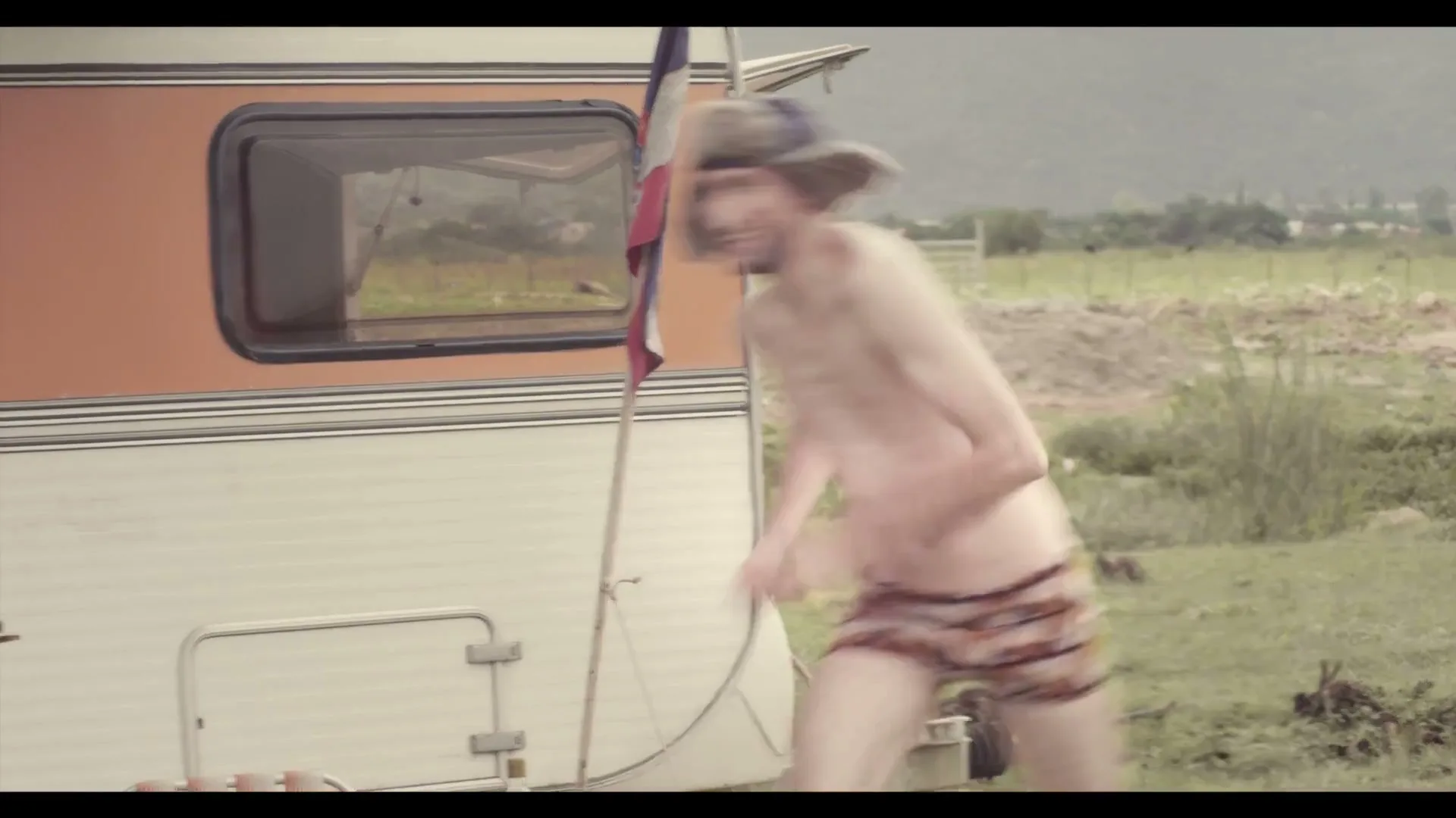 A shirtless man in a fur hat and animal print underwear runs past a camper van with a French flag in a rural, grassy field.