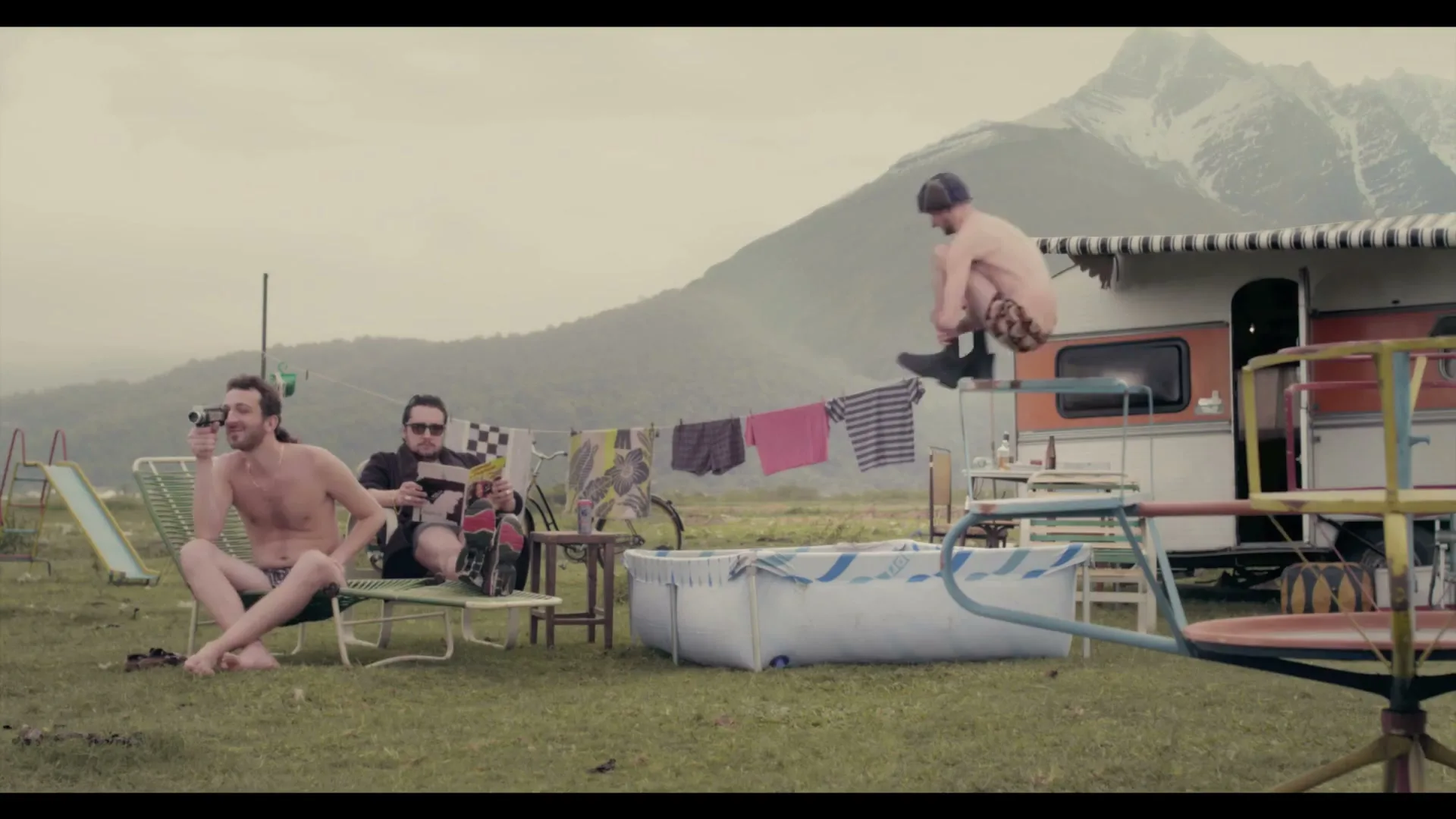 Three men are hanging out in a rural, mountainous area with a caravan and clothesline, as one man jumps off the caravan awning into a small pool.