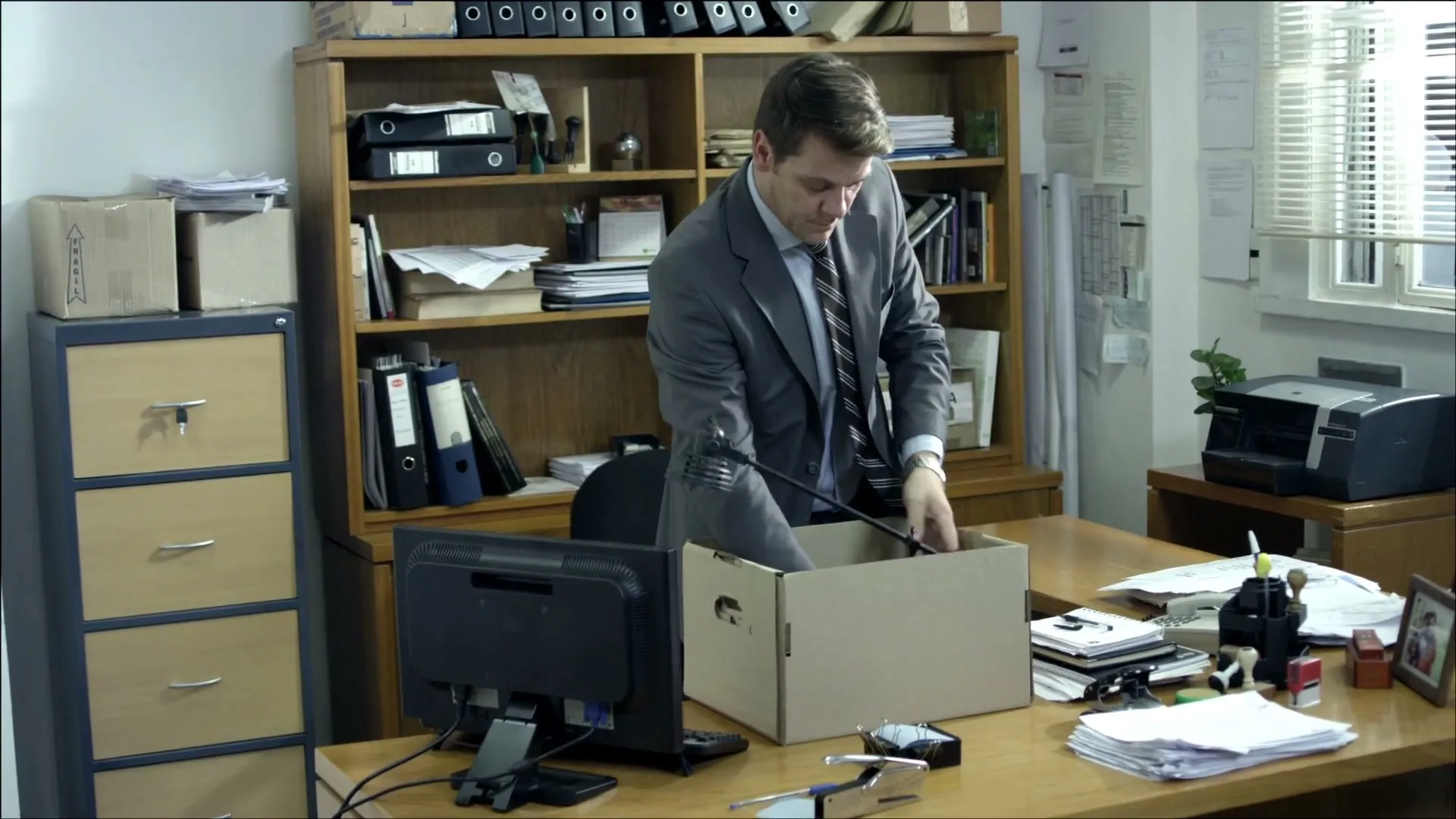 A man in a suit packs his belongings into a cardboard box on his desk in a cluttered office, looking sad.