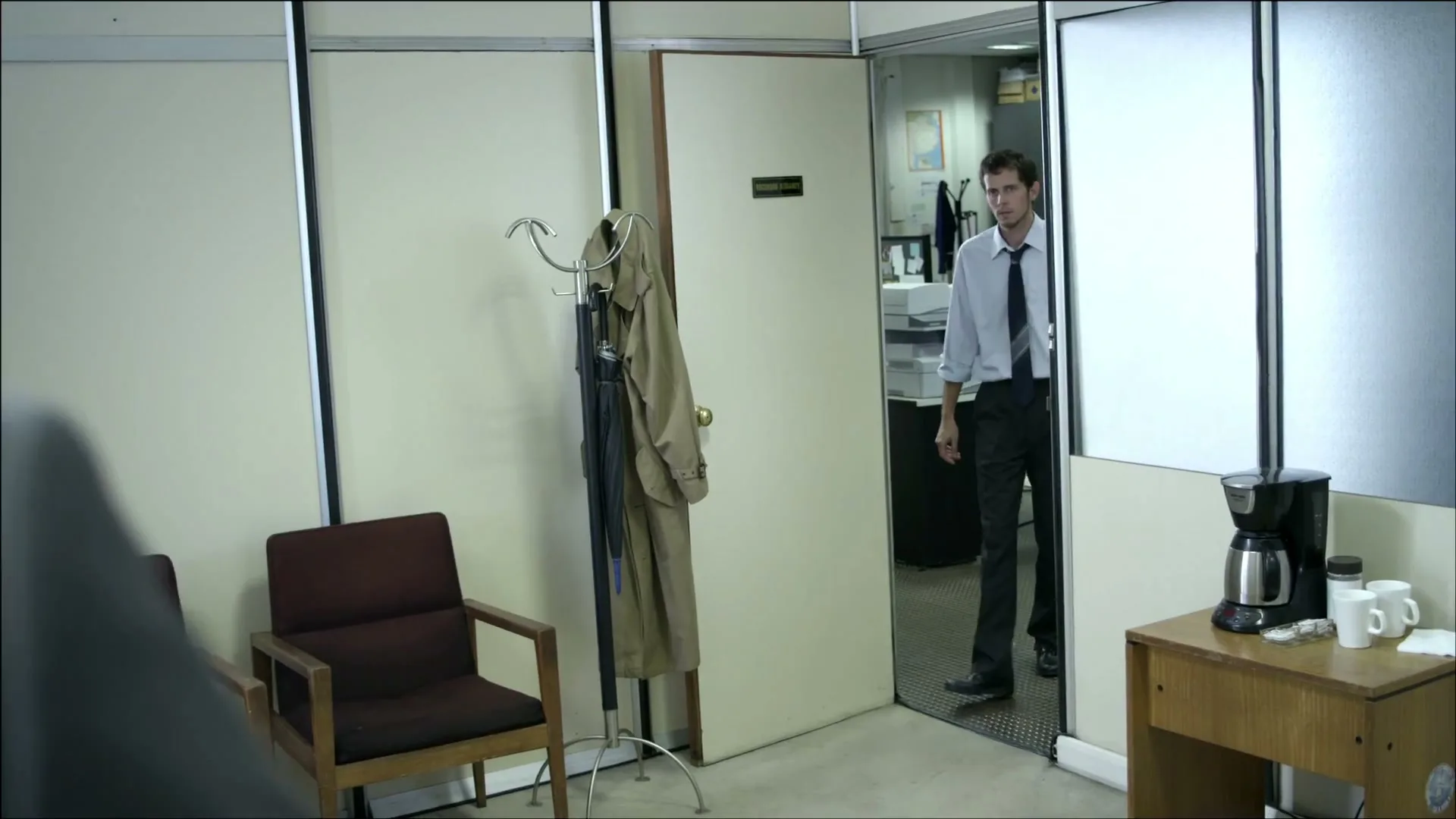A man in a dress shirt and tie stands in an office doorway, looking at someone off-screen, in a medium shot.