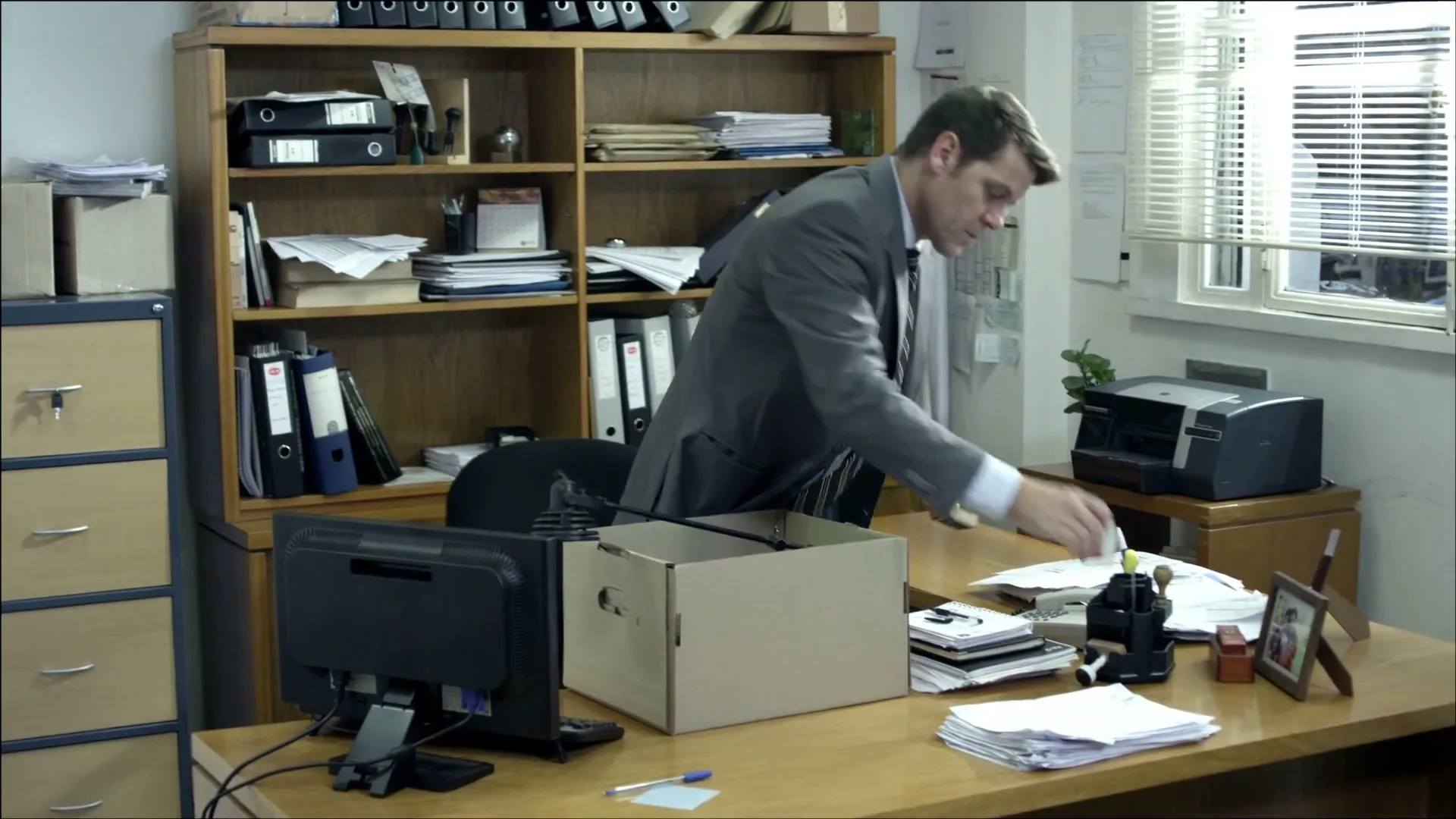 A man in a suit packs his belongings into a cardboard box on his desk in a cluttered office.
