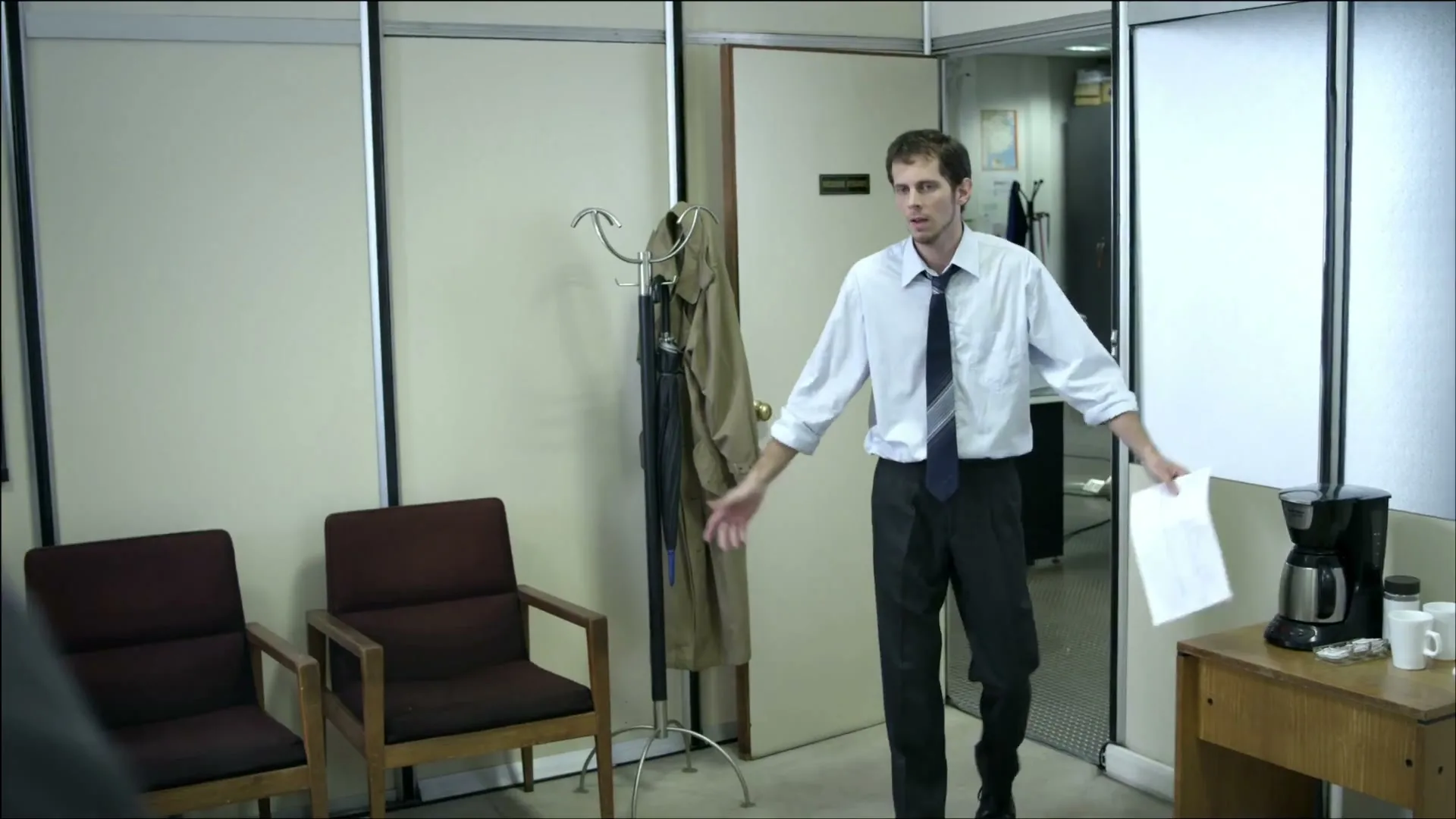 A man in a light blue shirt and dark tie walks into an office, holding papers and looking frustrated.