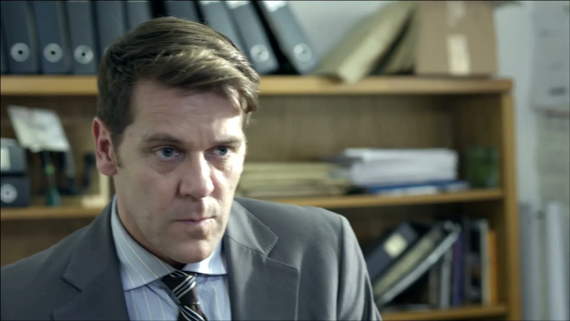 A man in a suit looks down, then up with a serious expression, in an office with shelves in the background.